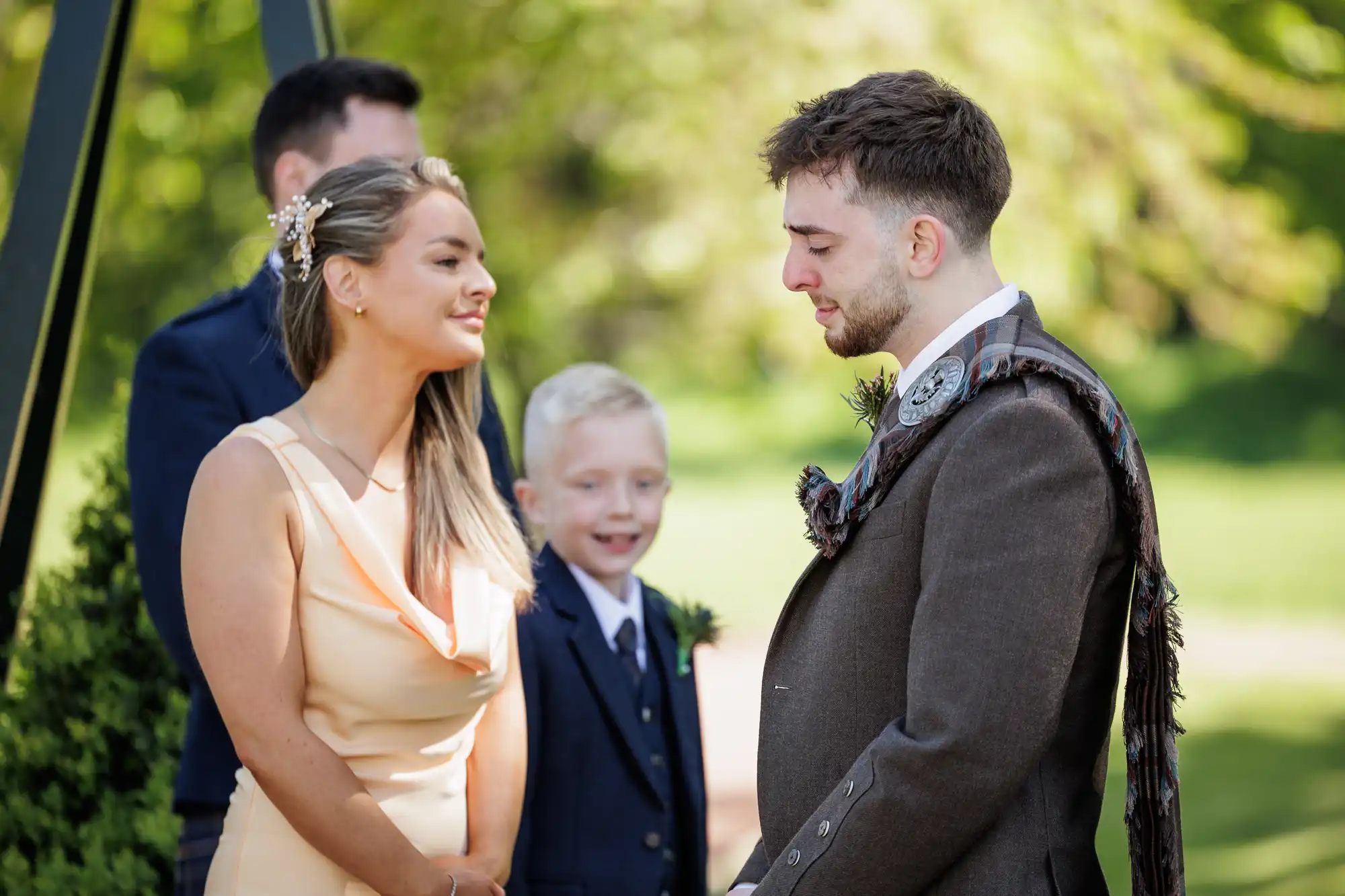 A woman in a peach dress and a man in brown Scottish attire stand facing each other outdoors, smiling. A young boy in a navy suit stands between them, smiling, with another man in a suit blurred in the background. Sunlit greenery surrounds them.