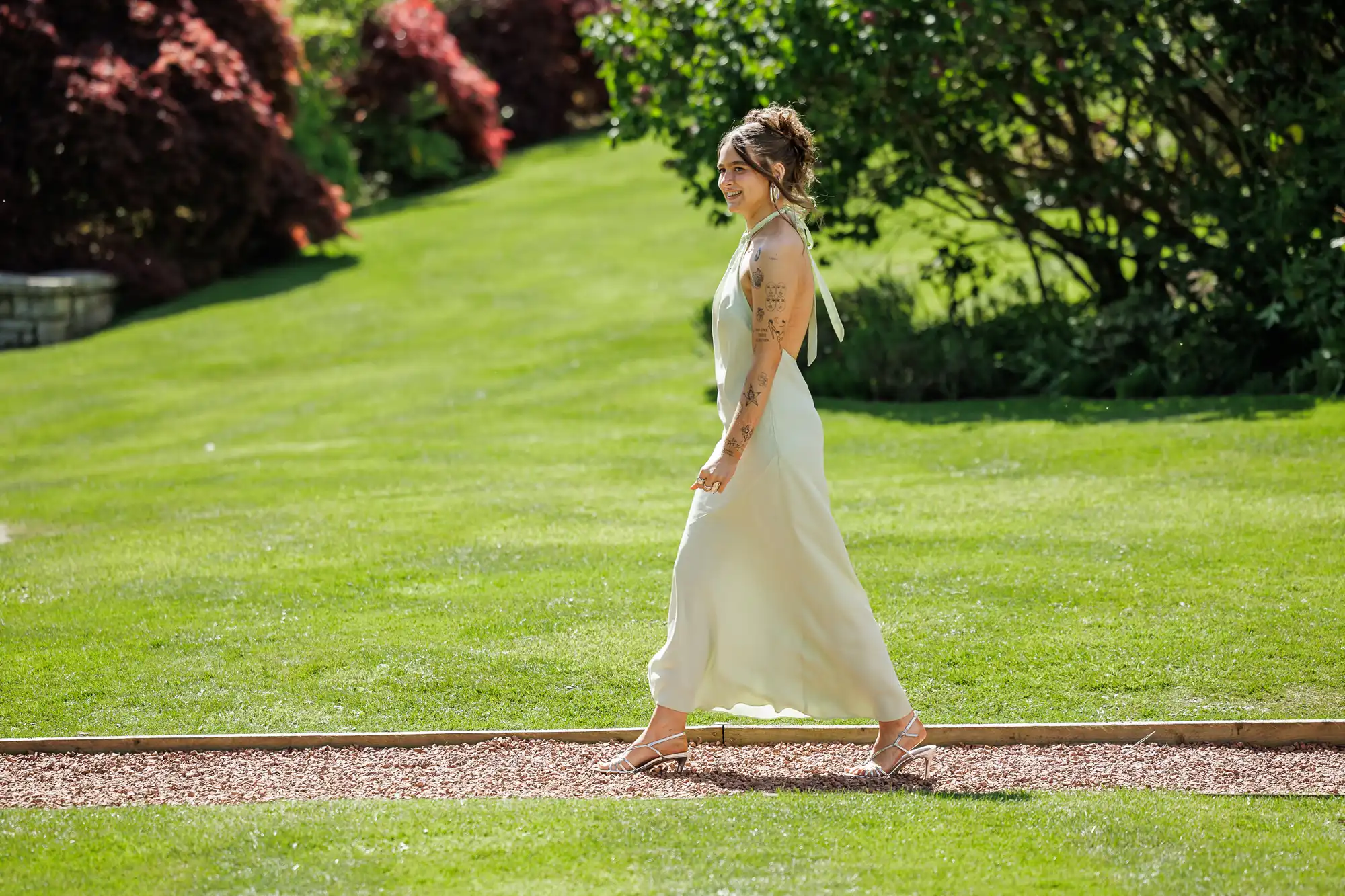 A woman in a long, light-coloured dress and sandals walks along a gravel path in a sunlit garden, surrounded by lush green grass and flowering bushes. She has tattoos on her arms and her hair is styled up.