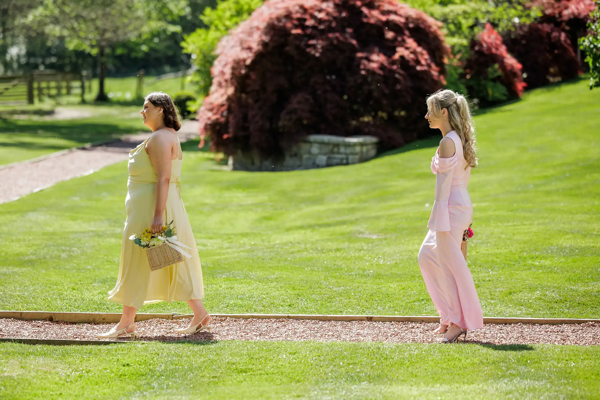Two women walk barefoot on a gravel path in a sunny, green garden. The woman in front wears a pale yellow dress and carries a basket of flowers. The woman behind wears a light pink dress and holds flowers behind her back. Shrubs and trees fill the background.