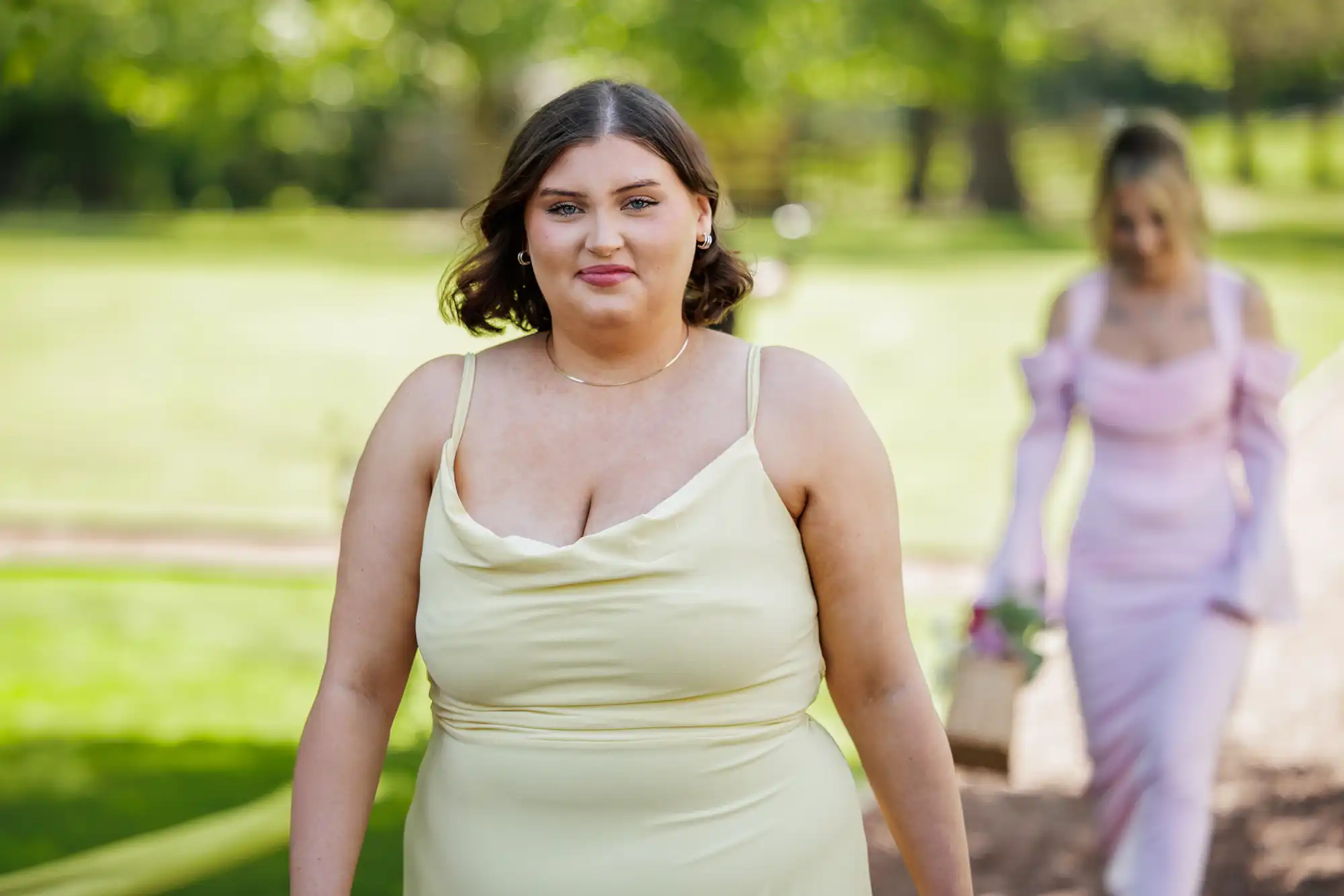 A woman with short brown hair in a pale yellow sleeveless dress walks outdoors on a sunny day, looking at the camera. Behind her, another woman in a light pink dress holds a basket and bouquet, slightly out of focus. Lush green trees and grass are in the background.