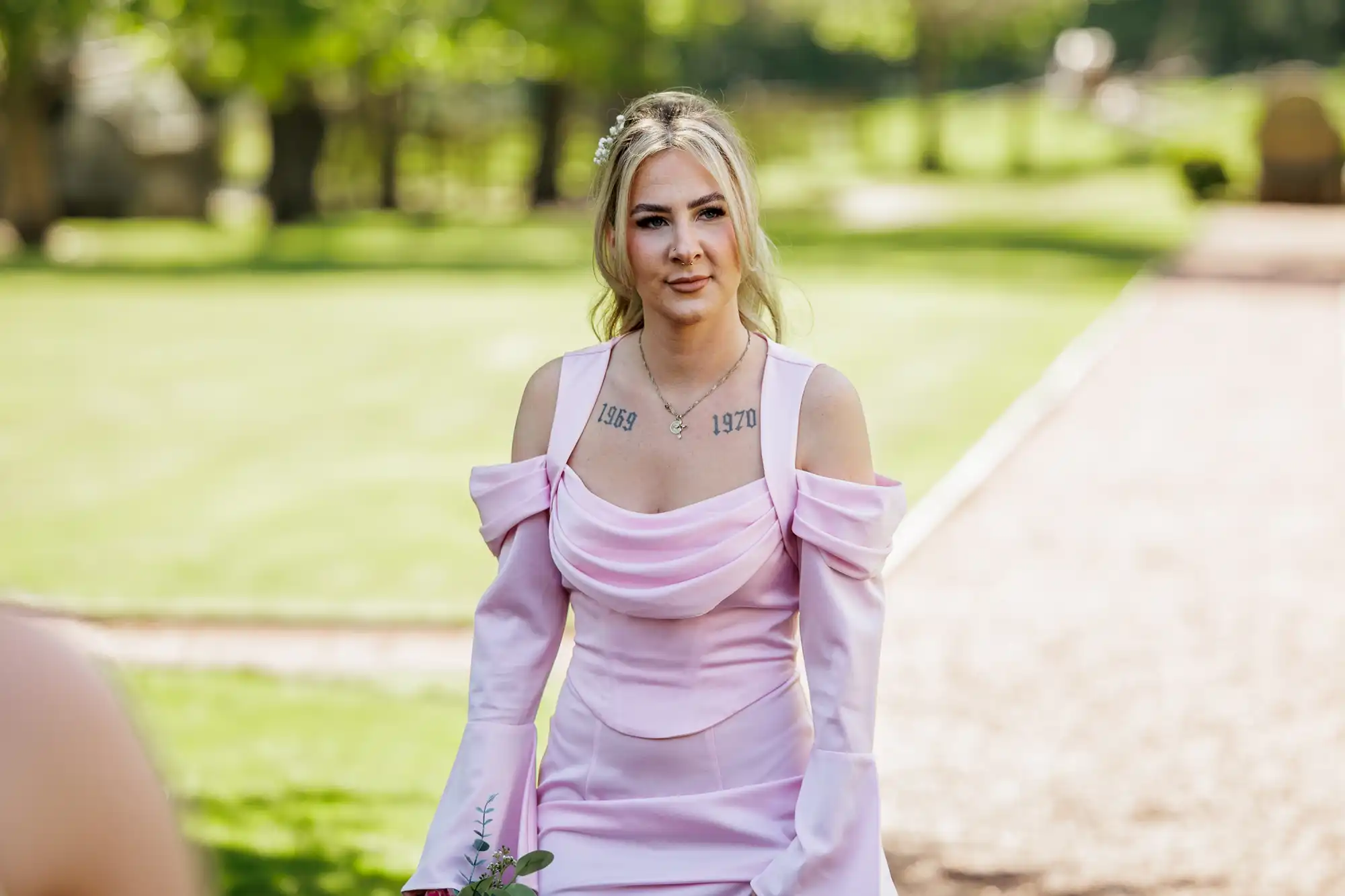 A woman with blonde hair in a half-up style stands outdoors on a sunny day, wearing a pale pink, off-the-shoulder dress. Visible tattoos with the numbers "1969" and "1970" are on her upper chest. Green trees and a lawn are blurred in the background.