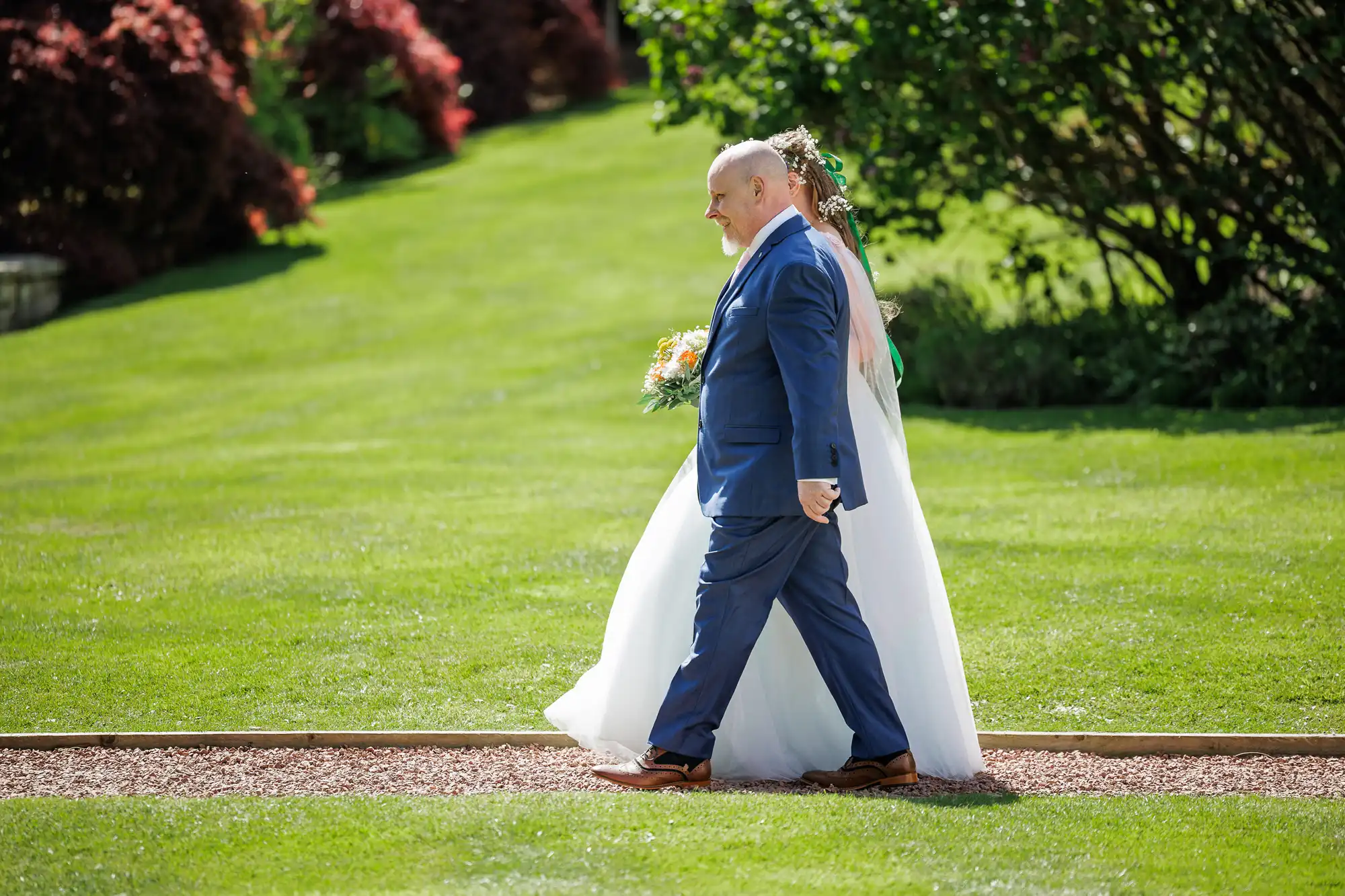 A bride in a white dress holding a bouquet walks outdoors on a grass path, accompanied by an older man in a blue suit. Lush green lawn and trees with red foliage are in the background, under bright sunlight.