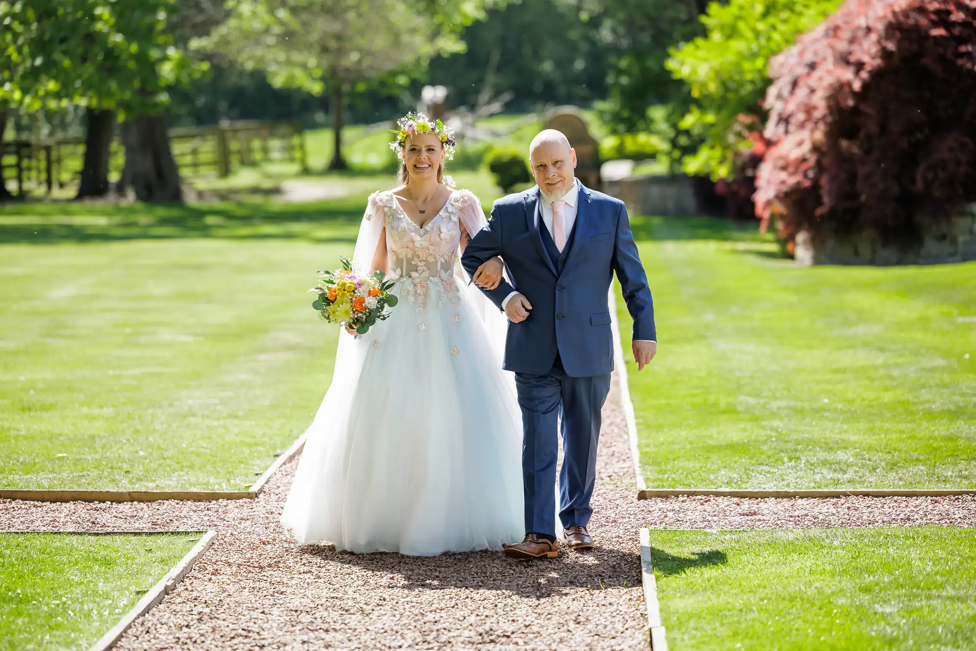 A bride in a white dress with floral details and a flower crown walks arm-in-arm with an older man in a blue suit and pink tie down a garden path on a sunny day. The bride holds a yellow bouquet; lush green grass and trees surround them.
