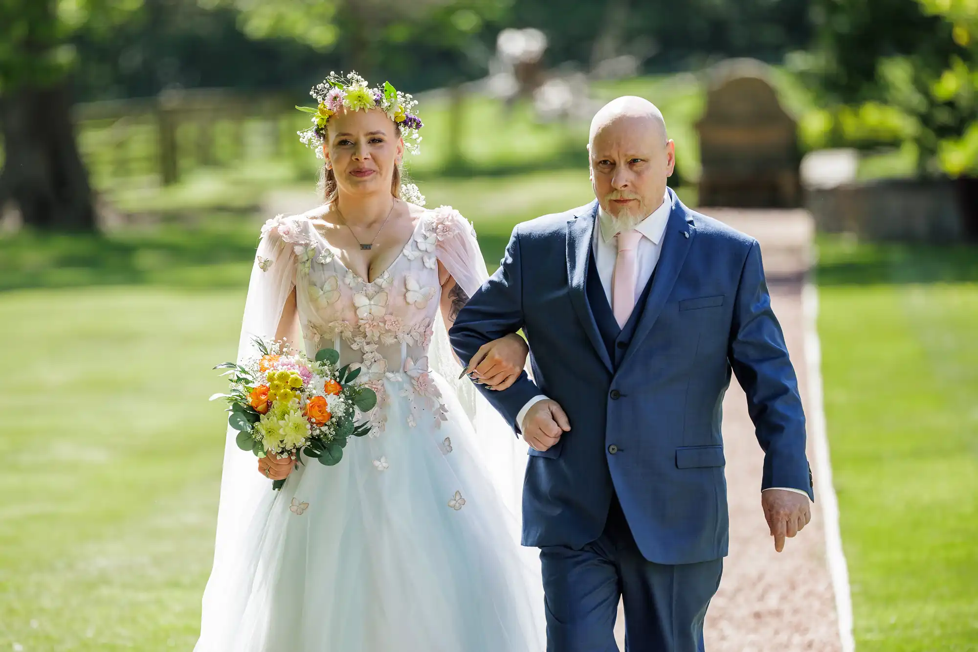 A bride in a white floral dress and flower crown holds a bouquet whilst walking arm-in-arm with a man in a blue suit and pink tie outdoors, down a garden path, with green grass and trees in the background.