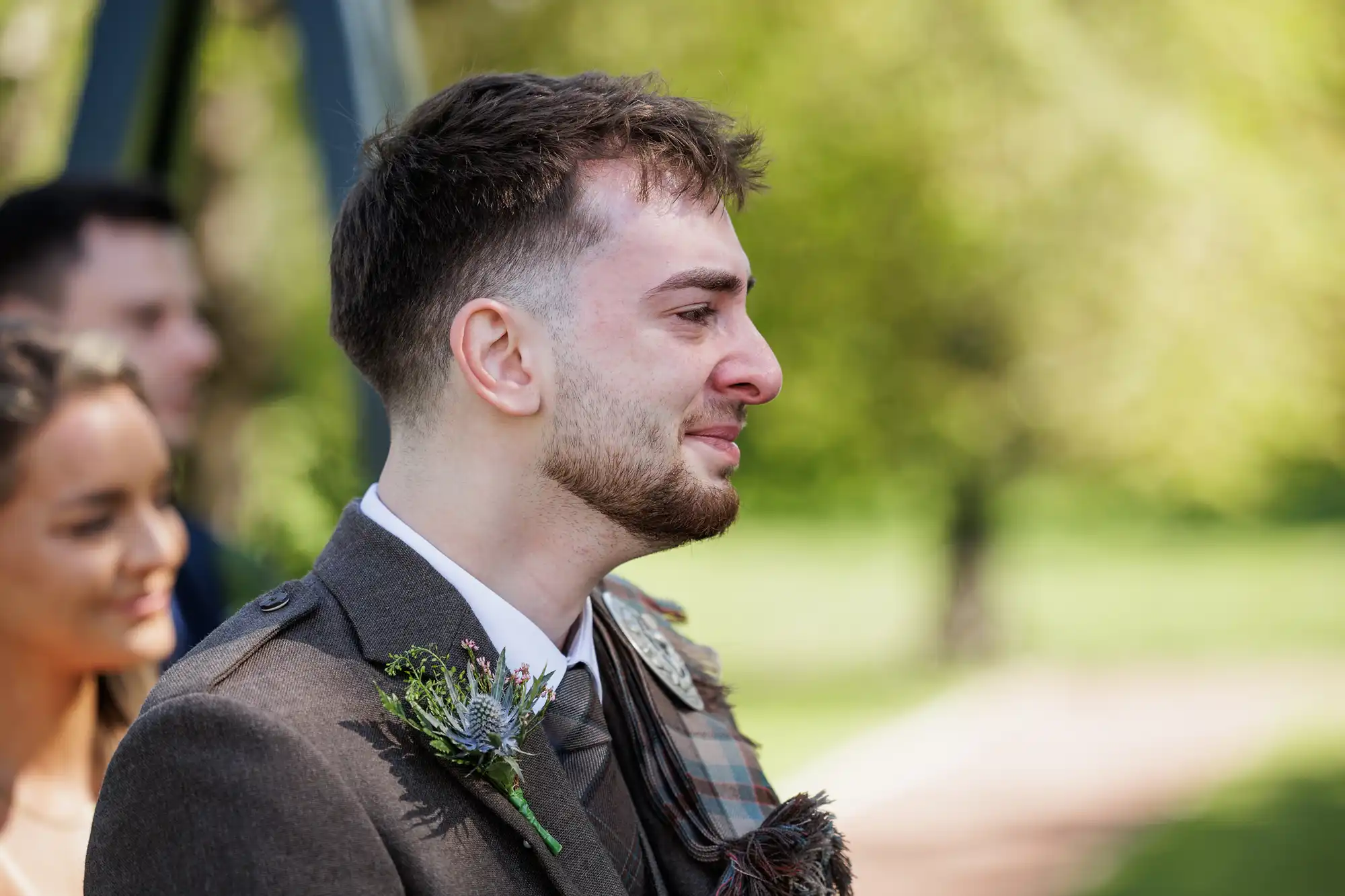 A young man wearing a brown suit with a tartan sash and buttonhole stands outdoors, appearing emotional with tears in his eyes. Behind him, a woman smiles softly. The background is blurred greenery and sunlight, suggesting a formal outdoor event.
