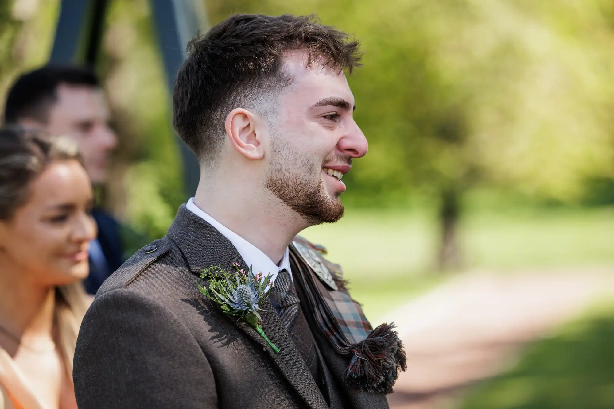 A smiling man wearing a brown suit jacket with a thistle buttonhole and tartan sash stands outdoors, with blurred greenery in the background. A woman with long hair stands behind him, slightly out of focus. It appears to be a formal or festive occasion.