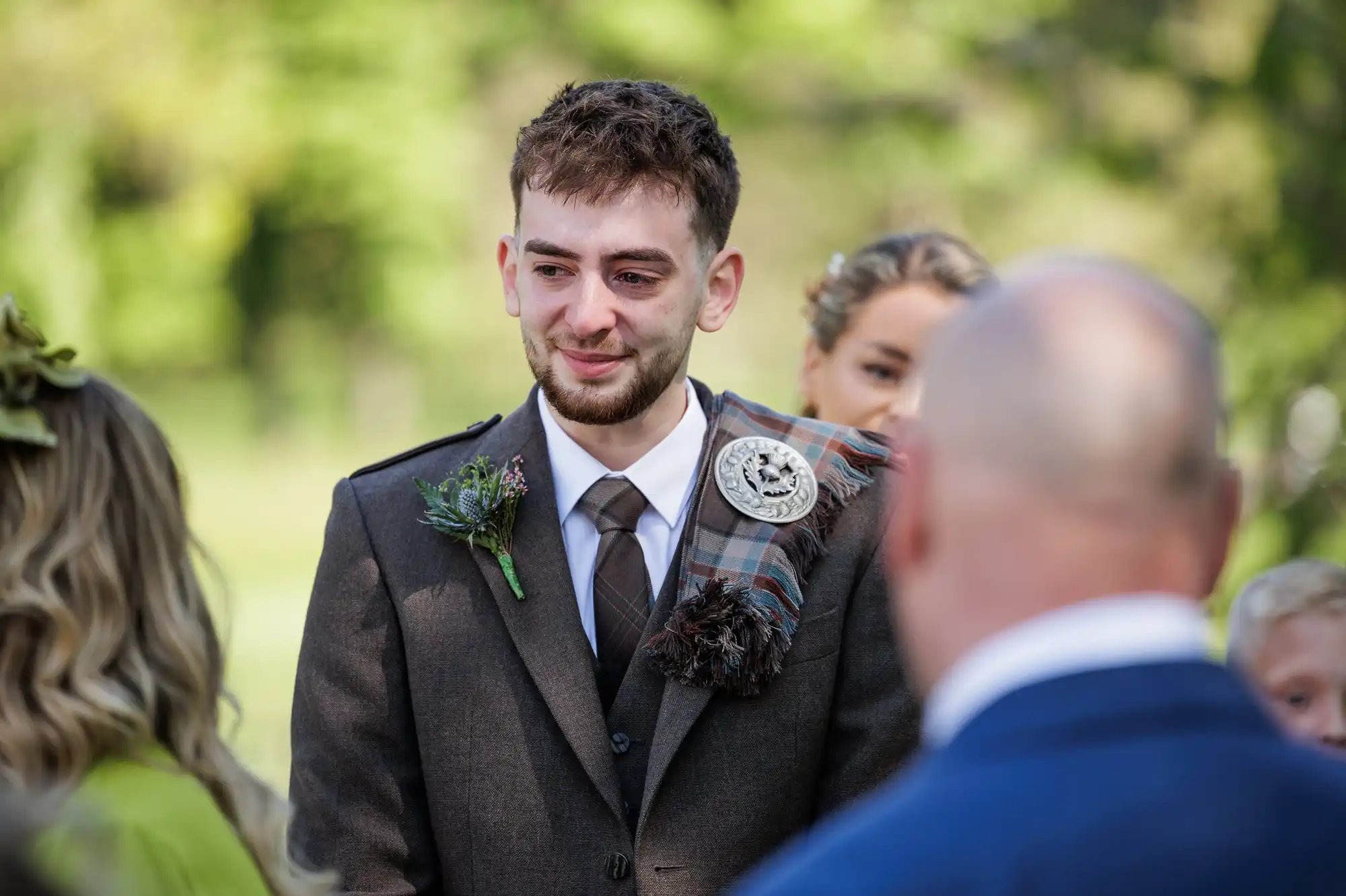 A young man in a brown suit with a checked sash and buttonhole smiles during an outdoor ceremony, facing a group of people. The background is green and blurred, suggesting a garden setting. Others, partially visible, stand around him.
