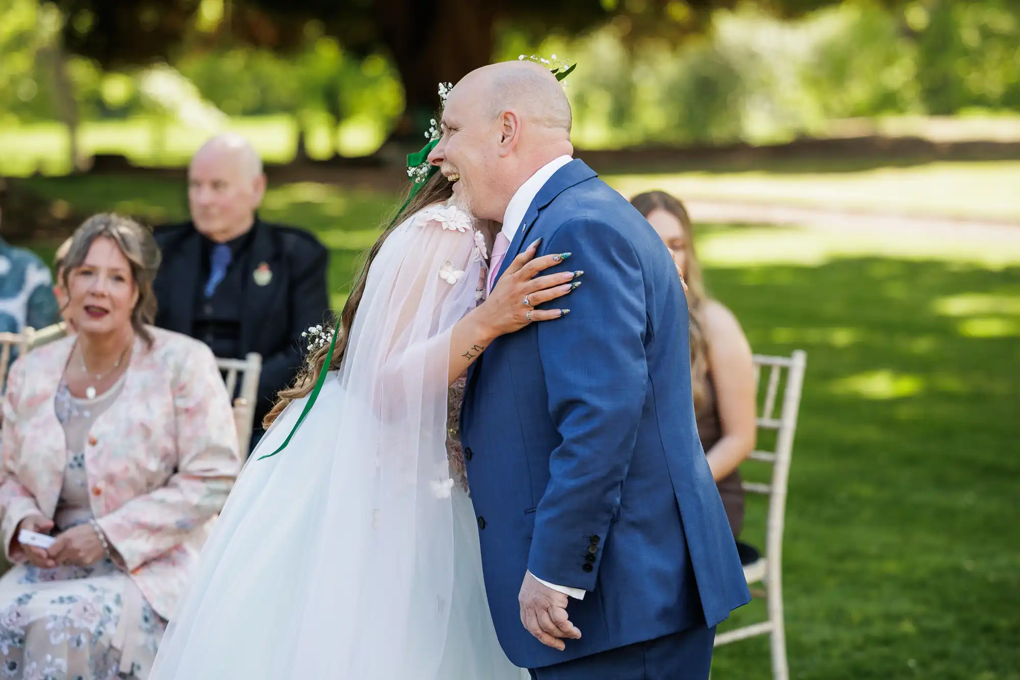 A bride in a white dress and veil hugs a bald man in a blue suit outdoors. Guests sit on white chairs, including a woman in a floral jacket. Green grass and trees provide a sunny, blurred background.