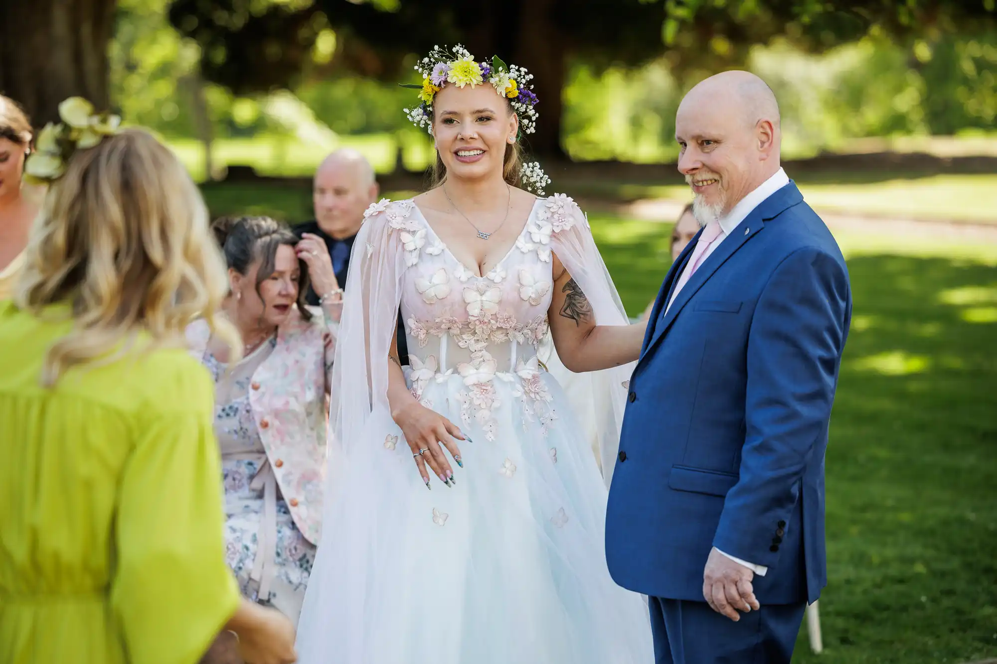 A smiling bride in a white floral gown and flower crown stands beside a man in a blue suit. They are outdoors, surrounded by greenery, with guests seated behind them. Sunlight filters through the trees, creating a festive, joyful atmosphere.