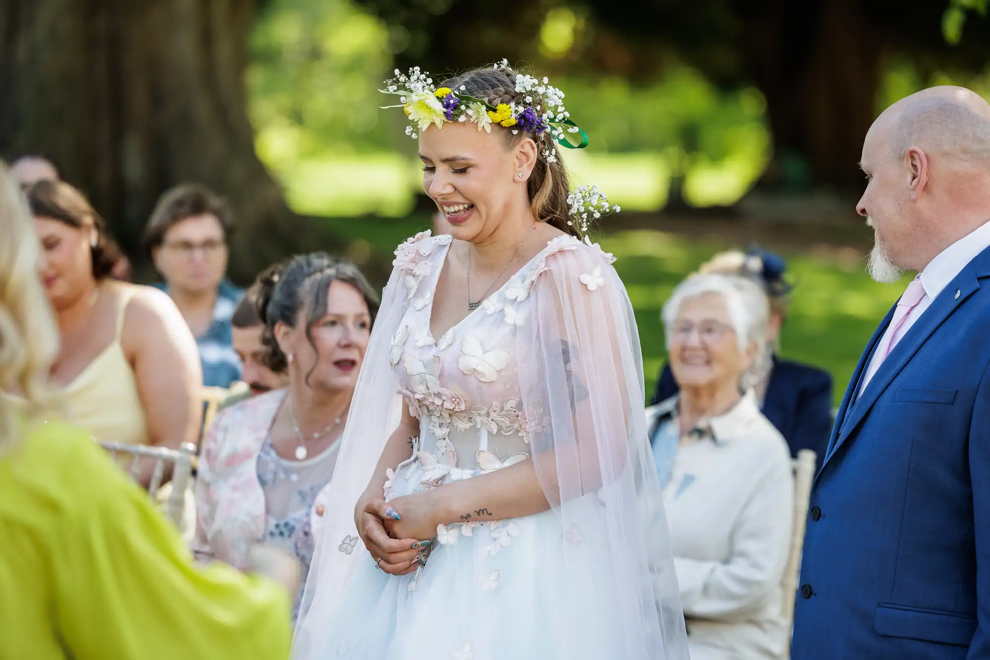 A smiling bride in a floral crown and white dress stands outdoors among seated guests. She wears a cape with flower appliqués. Sunlight filters through trees behind her. An older man in a blue suit and pink tie stands nearby, facing her. Everyone appears happy and relaxed.