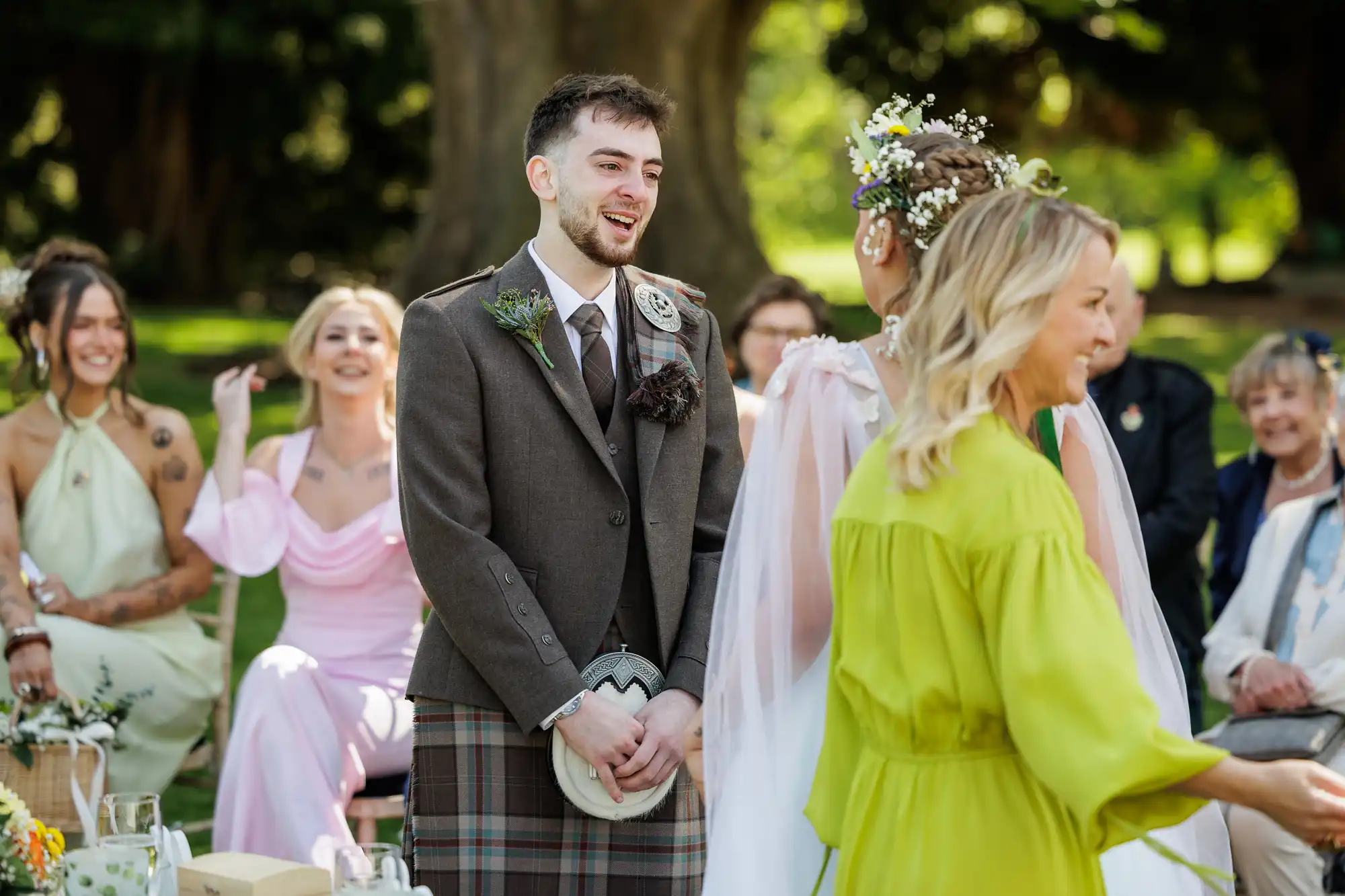 A groom in a brown jacket and tartan kilt smiles at a bride wearing a white dress and floral headpiece during an outdoor wedding ceremony. Guests sit nearby; one woman in the foreground wears a bright yellow dress. Sunlight filters through green trees in the background.