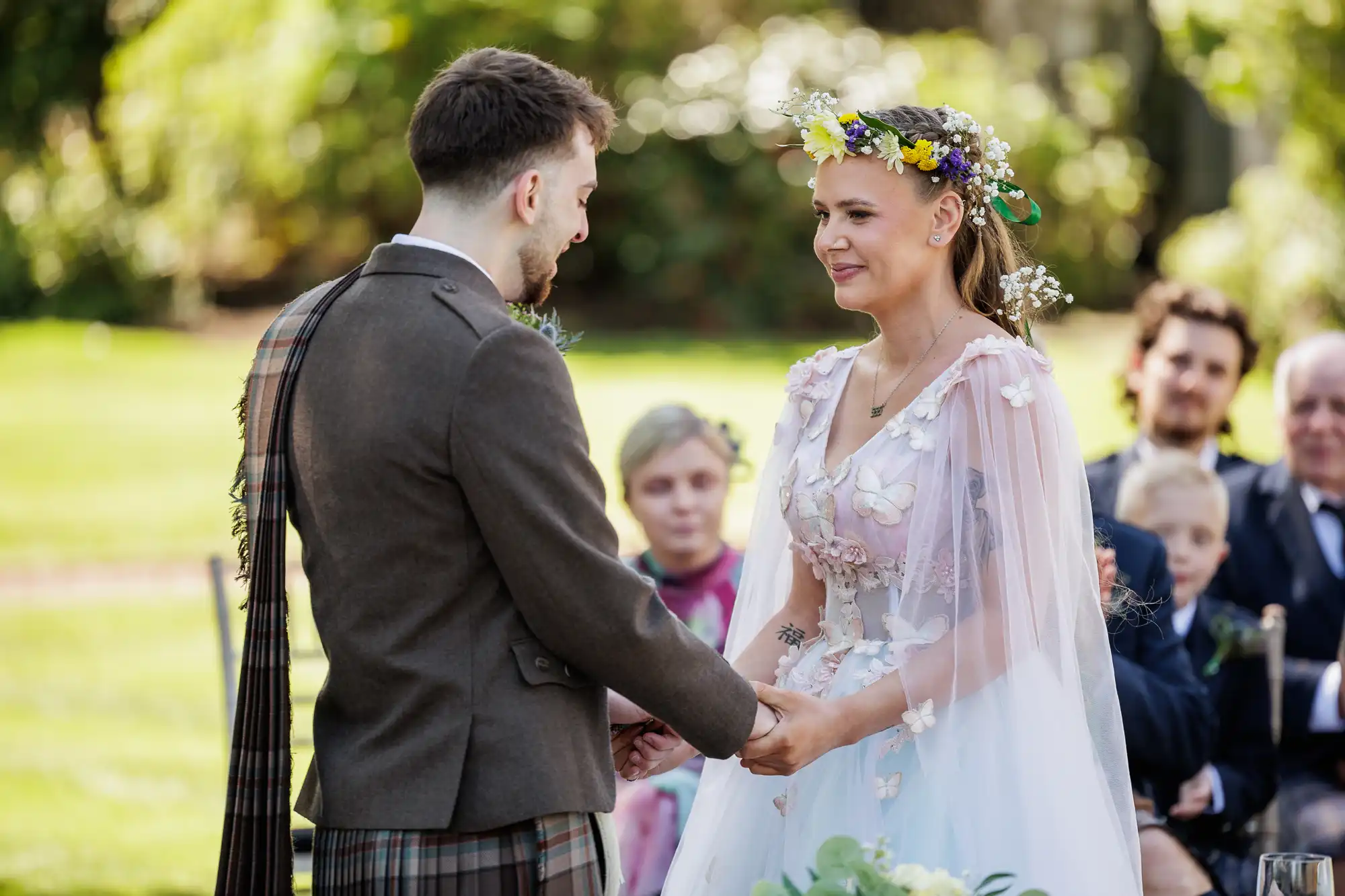 A bride and groom stand holding hands outdoors during their wedding ceremony. The bride wears a floral crown and a pastel dress with flower details, whilst the groom wears a brown jacket and tartan sash. Guests are seated and watching in the background.