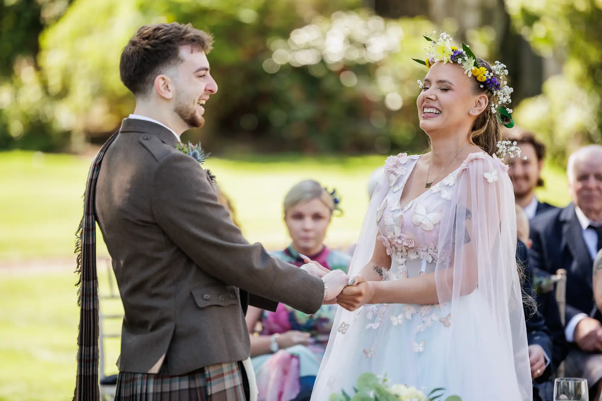 A couple stands outdoors, holding hands and smiling joyfully at each other during their wedding ceremony. The bride wears a flower crown and a dress with floral accents; the groom wears a suit with a kilt. Guests are seated in the background, watching.