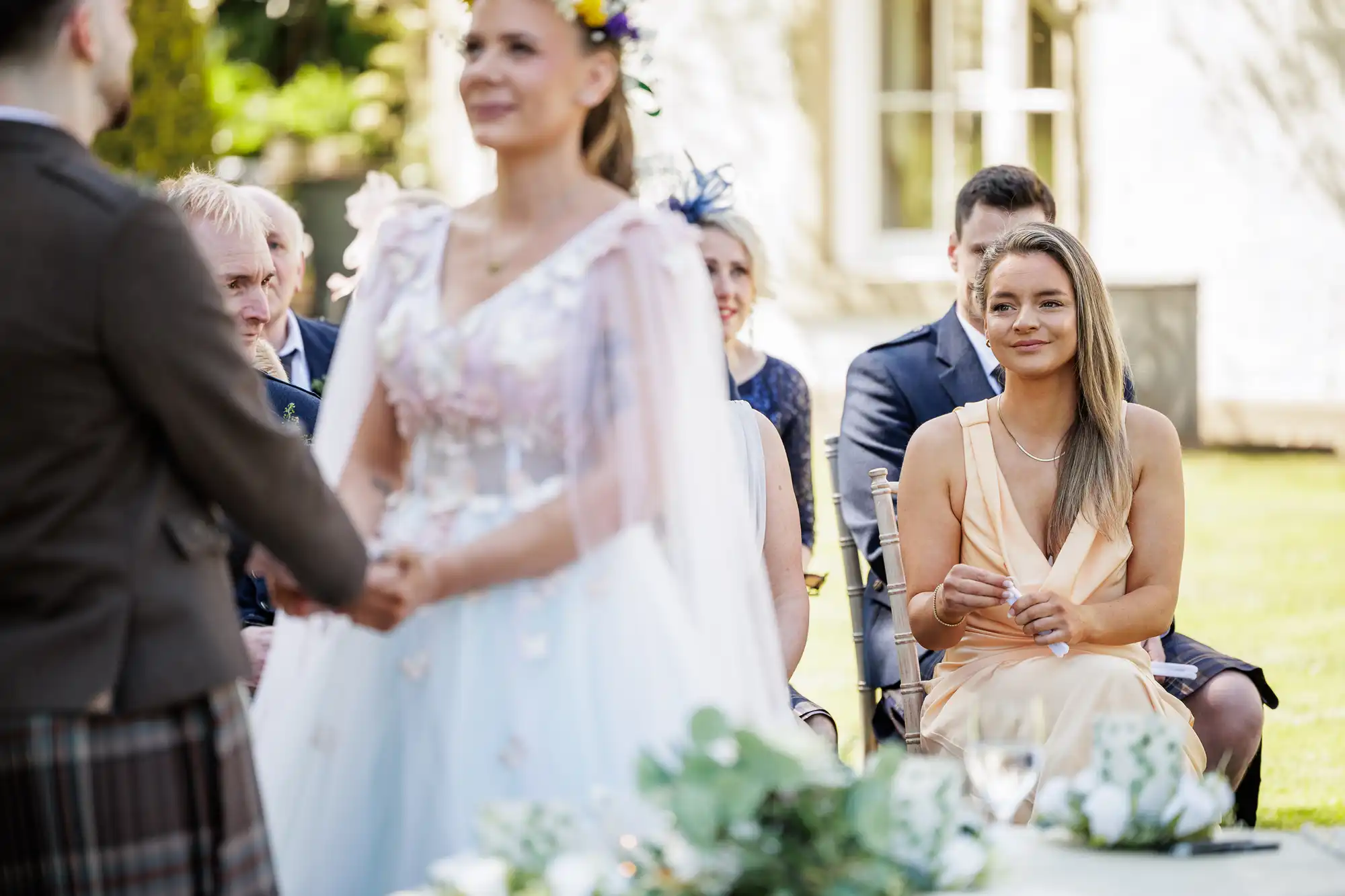 A bride and groom hold hands during an outdoor wedding ceremony. The bride wears a pastel floral dress and veil. Guests, including a woman in a pale dress smiling, are seated, with greenery and sunlight all around. The atmosphere is joyful and elegant.