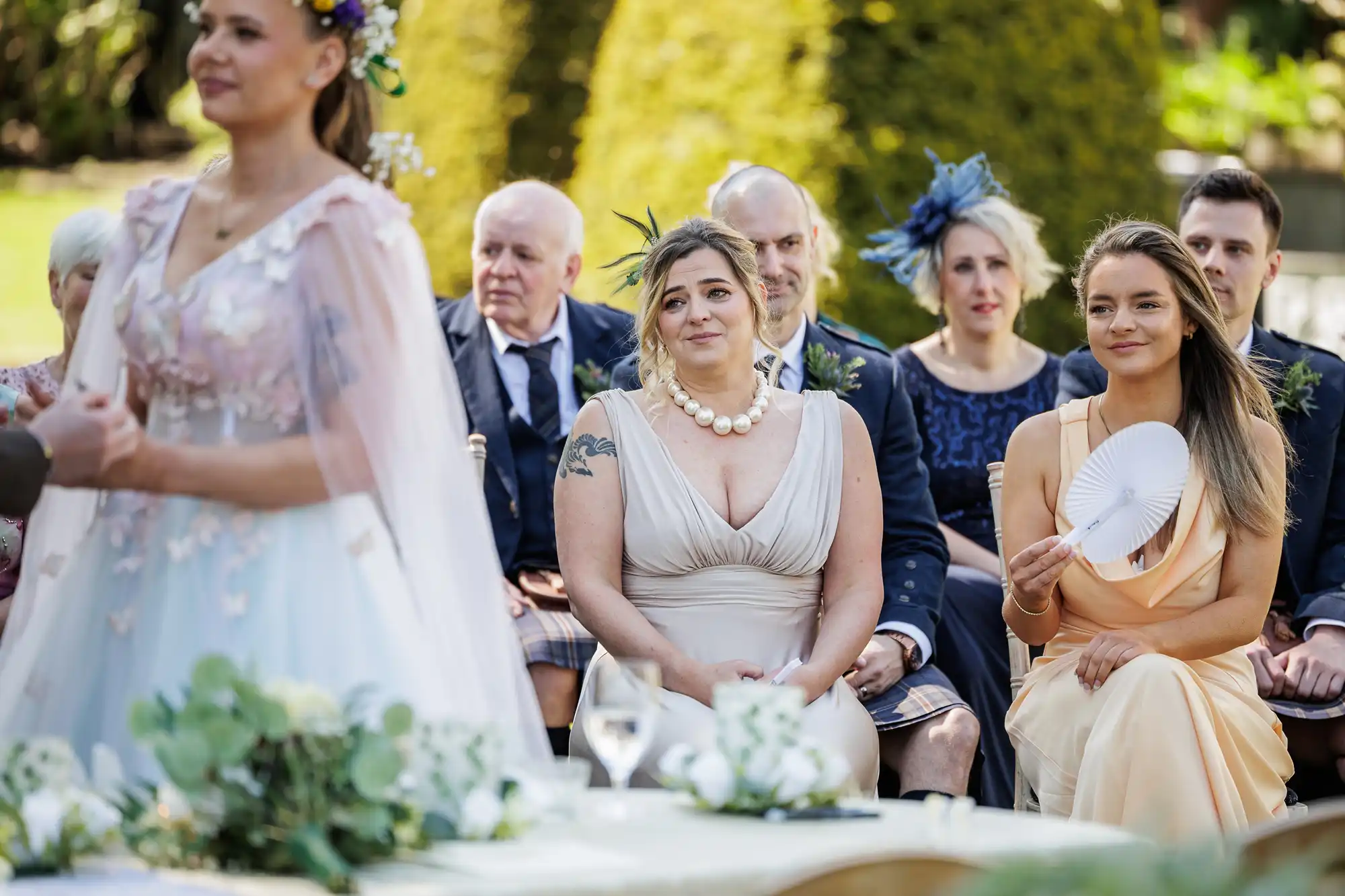 A group of wedding guests sit outdoors, watching a ceremony. In the foreground, two women in formal dresses, one in light beige with a pearl necklace and one in pale yellow holding a fan, are seated. Greenery and blurred people appear in the background.