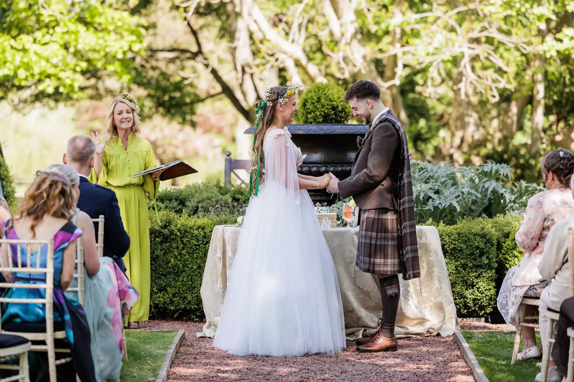 A bride in a white dress and flower crown holds hands with a groom in a kilt during an outdoor wedding ceremony. Guests sit on either side, and an officiant in a yellow dress stands behind them, smiling. Green trees and sunlight fill the background.