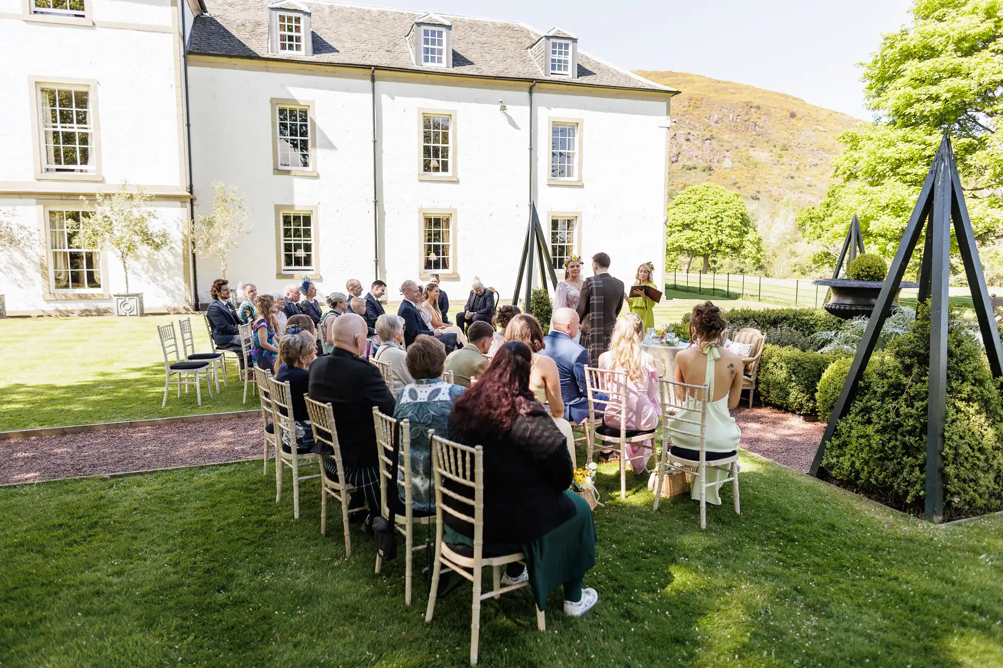 A group of people sit on white chairs arranged in rows on a lawn, attending an outdoor event by a large white building on a sunny day. The setting is surrounded by greenery, with mountains and trees visible in the background.