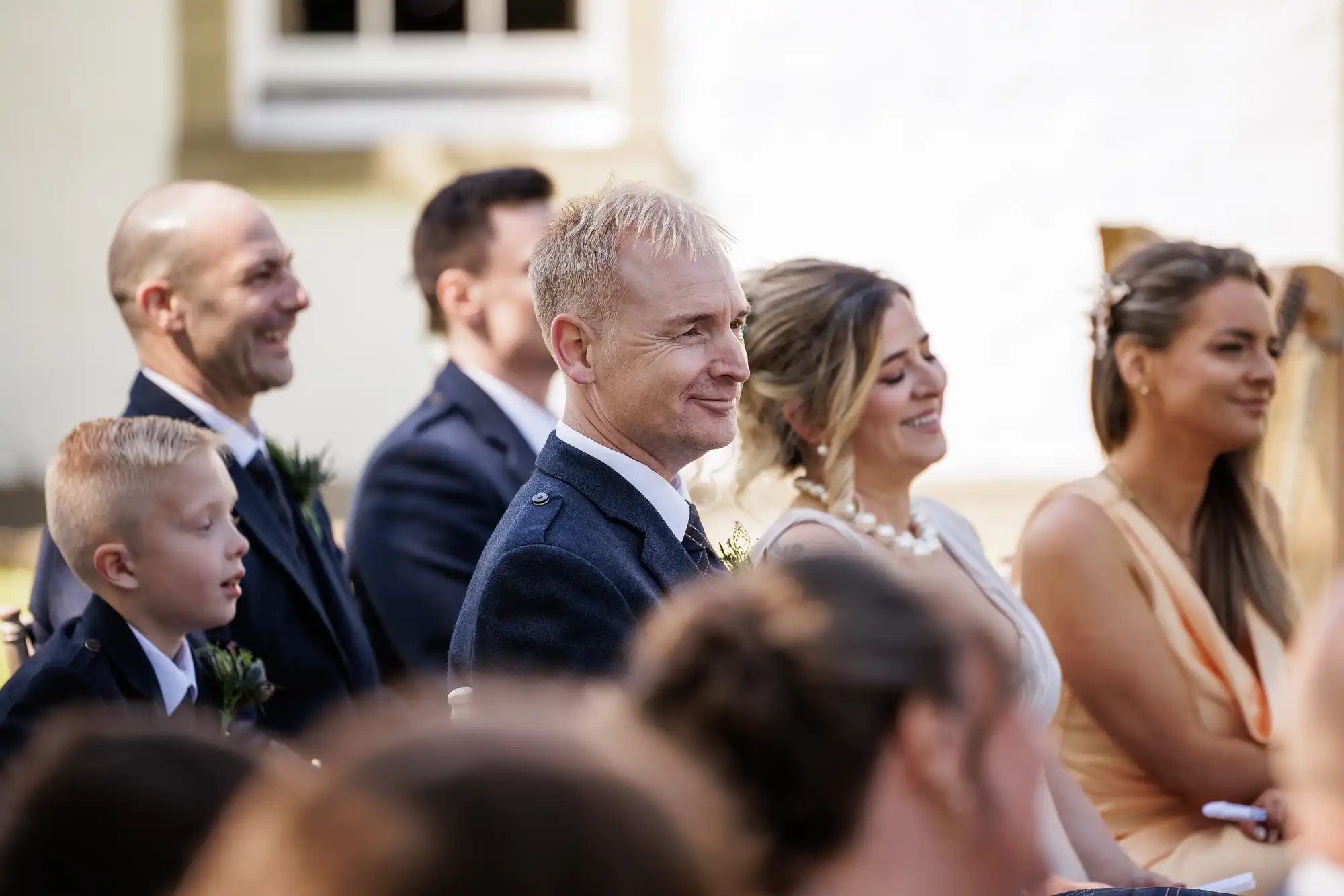A group of well-dressed people, including men in suits and women in formal dresses, sit closely together outdoors at a wedding ceremony, smiling and looking forwards. Sunlight brightens the scene, and a building is visible in the background.