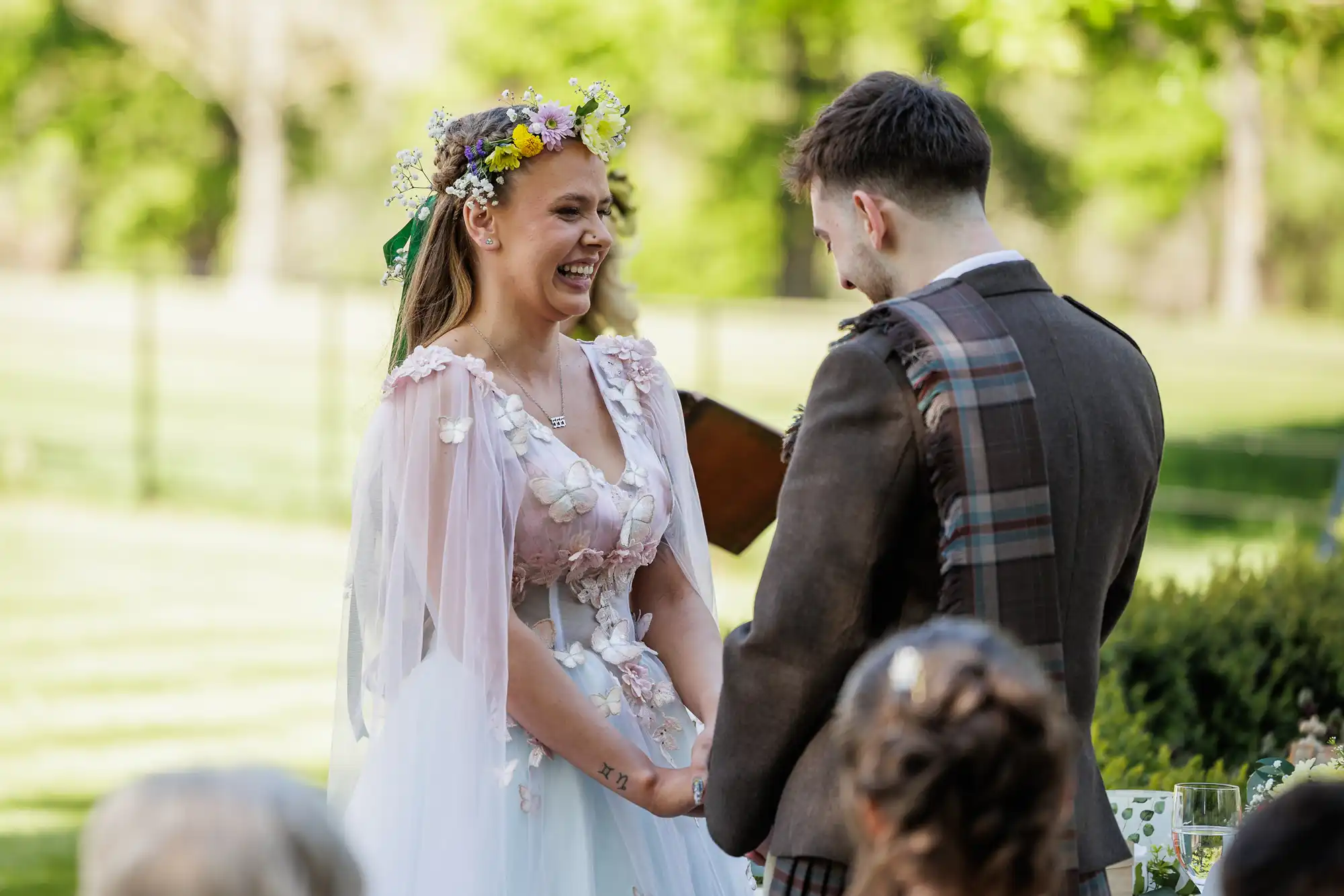 A bride in a floral headpiece and pastel dress smiles joyfully, holding hands with a groom in a brown suit with tartan check accents, outdoors on a sunny day, surrounded by greenery and guests watching the wedding ceremony.