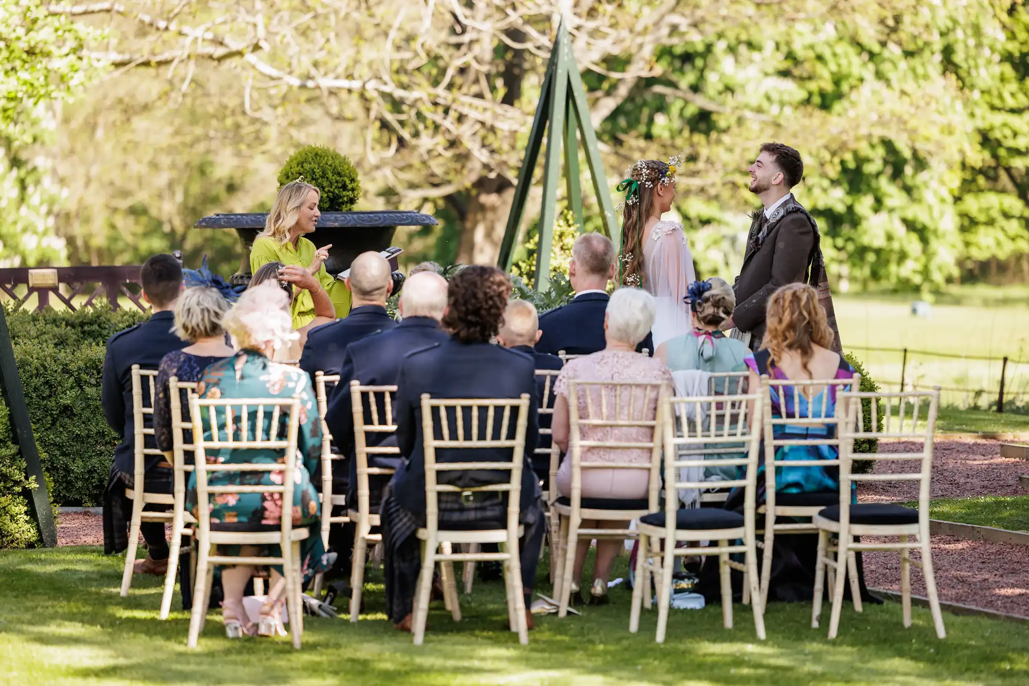 A small outdoor wedding ceremony with guests seated on wooden chairs facing a couple standing before an officiant. The bride wears a white dress and flower crown, the groom a dark suit. Trees and greenery create a bright, sunny background.