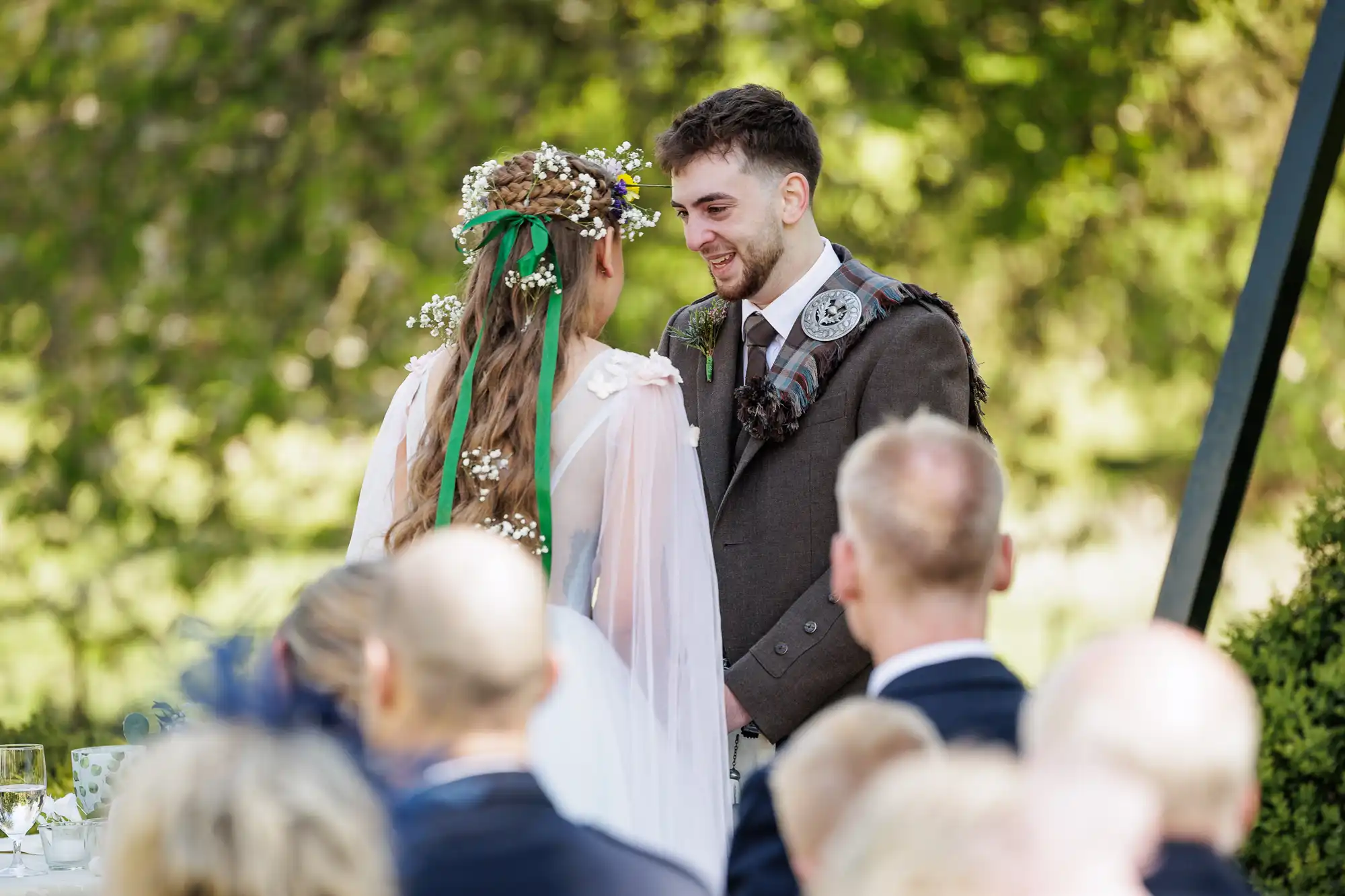 A bride and groom face each other outdoors, smiling during their wedding ceremony. The bride wears a white dress and floral crown; the groom wears a brown suit with a checked sash. Guests, blurred in the foreground, watch as trees and greenery fill the background.