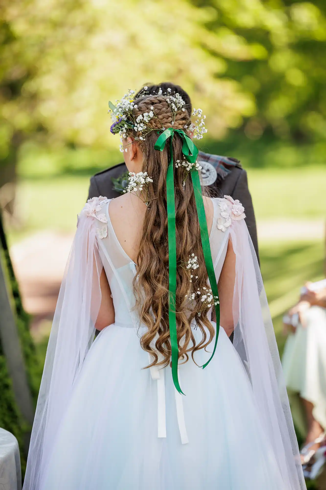 A bride seen from behind wears a white dress with sheer sleeves, long wavy hair adorned with flowers and green ribbons, and a floral crown. She stands outdoors in sunlight, facing a person in a suit, with greenery blurred in the background.