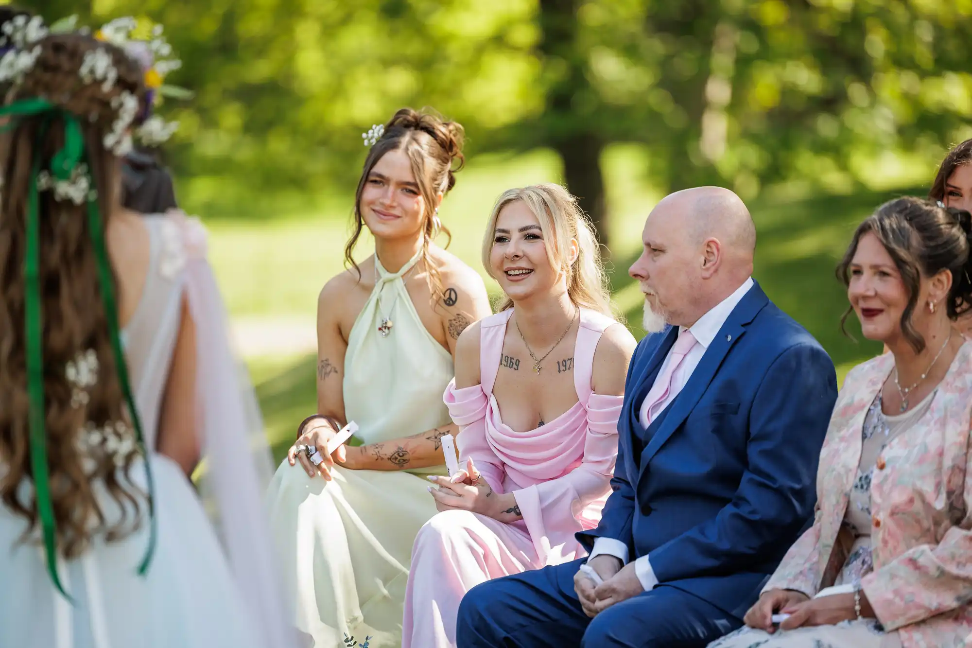 Three guests—two women in pastel dresses and a man in a blue suit—sit outdoors, smiling at a bride in a white gown. The background is lush and green, suggesting a sunny, joyful wedding ceremony in a park or garden setting.