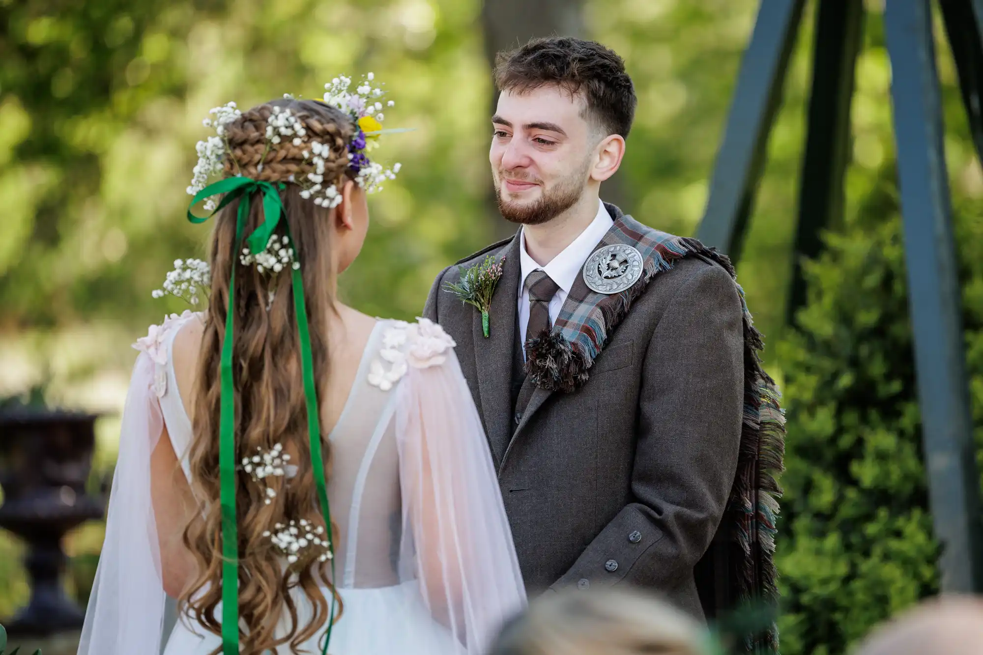 A groom in a brown suit with a tartan sash smiles at a bride wearing a white dress with sheer sleeves and flowers in her long hair. They stand outdoors, facing each other, surrounded by greenery and soft sunlight.