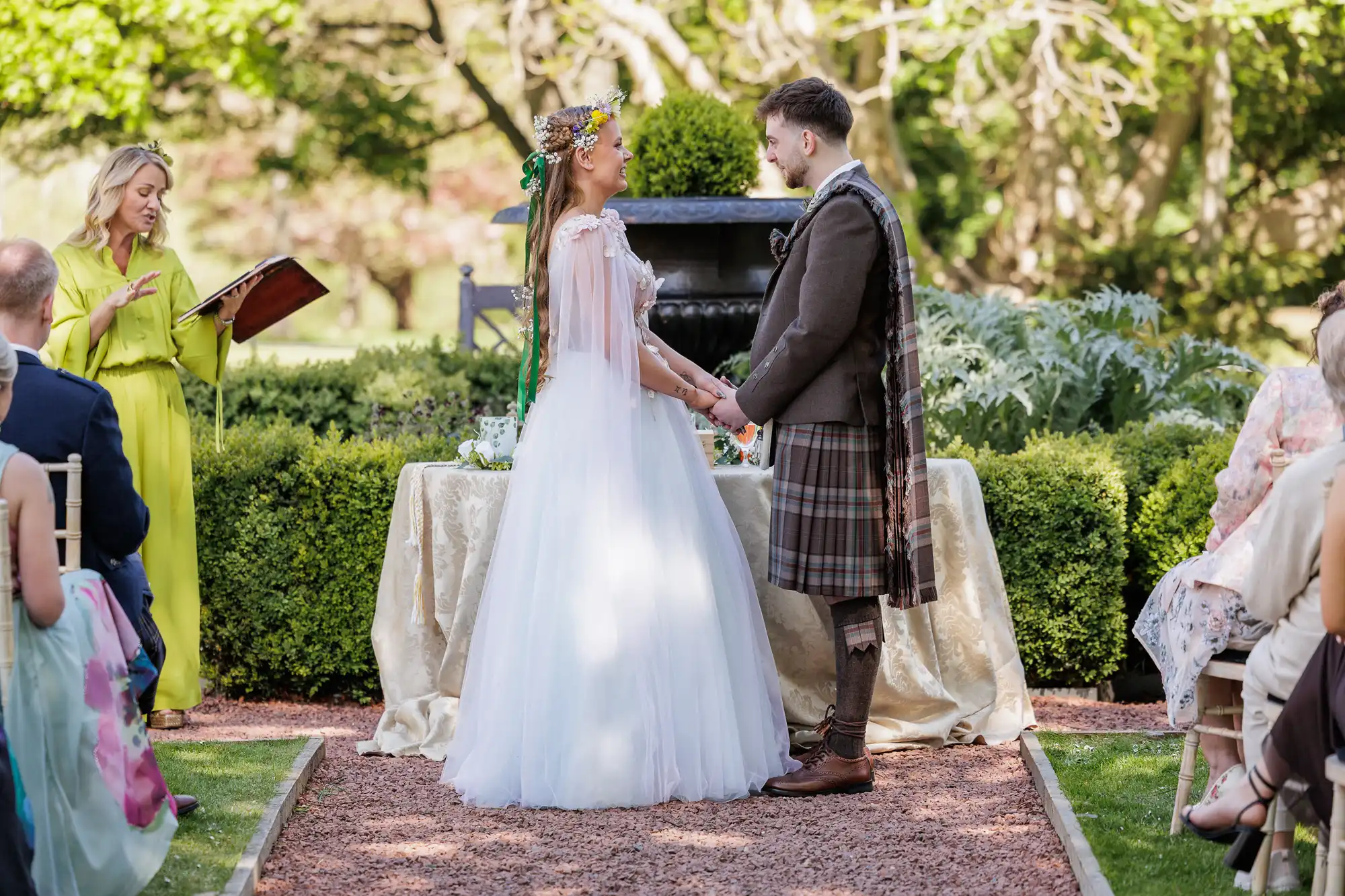A bride in a white gown and floral crown holds hands with a groom in a brown jacket and tartan kilt at an outdoor wedding ceremony. An officiant in a bright yellow dress reads from a book as seated guests watch. Lush greenery and sunlight fill the background.
