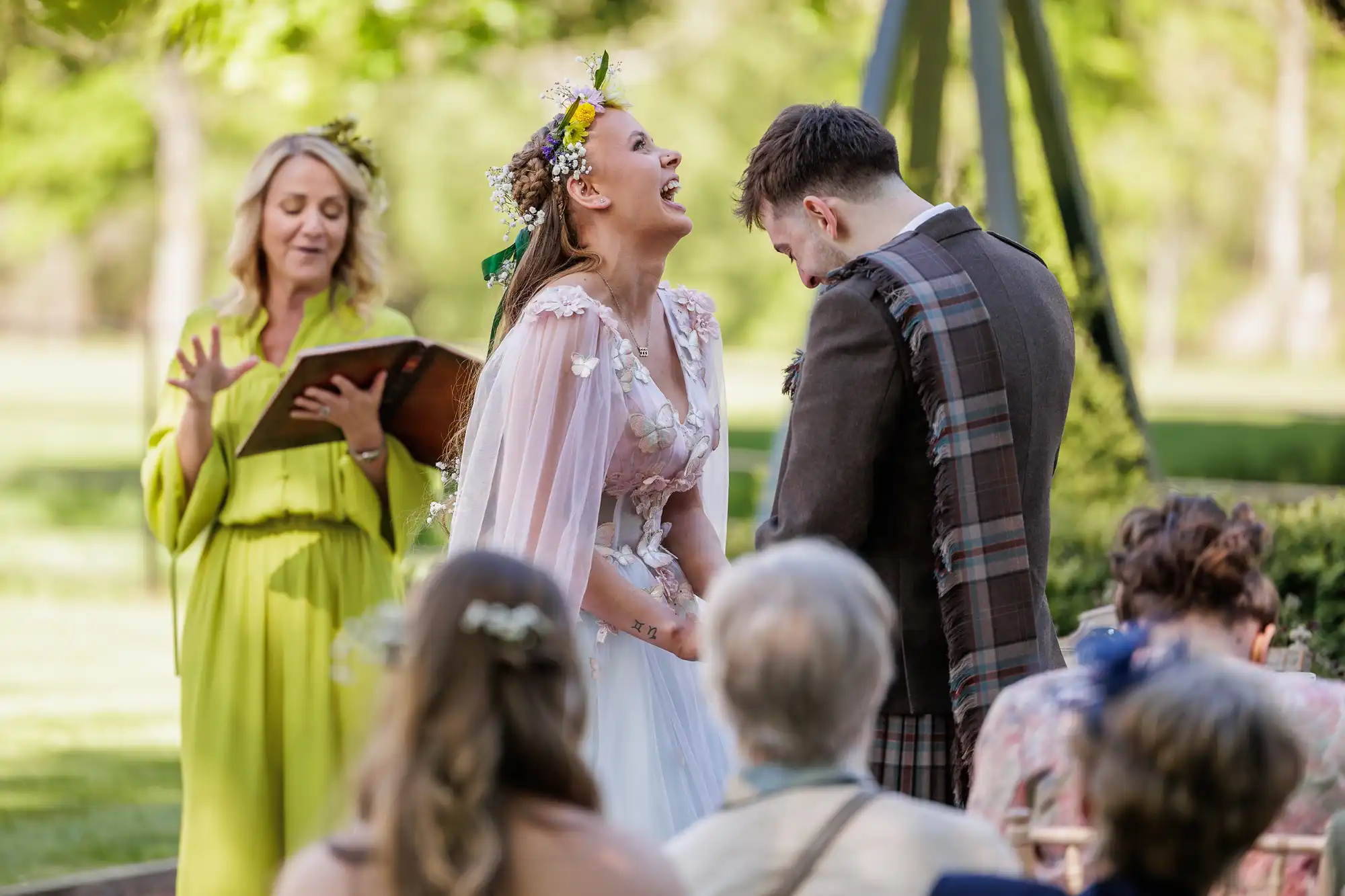 A bride in a floral crown and pastel dress laughs joyfully beside a groom in a checked sash during an outdoor ceremony. An officiant in a bright yellow dress reads from a book. Seated guests watch, surrounded by greenery and sunlight.
