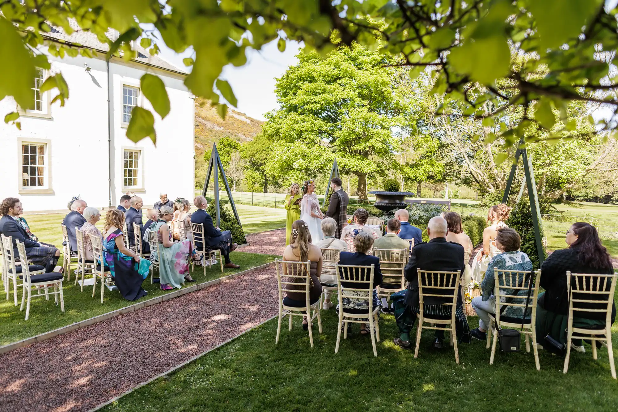 A small outdoor wedding ceremony with guests seated on white chairs facing a couple standing at the altar. Green trees and grass surround the scene, and a white building stands on the left under a sunny sky with leafy branches framing the top of the image.