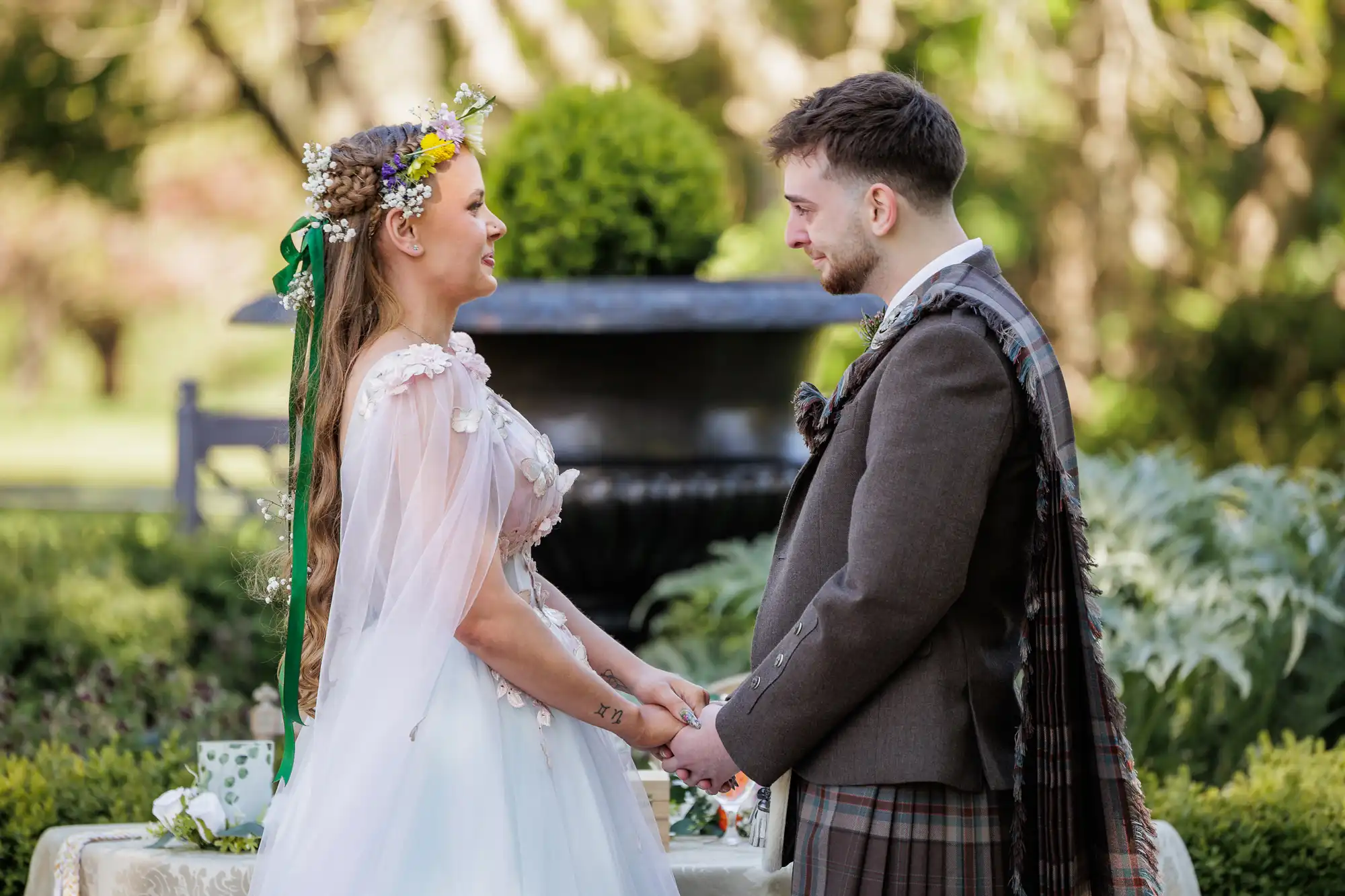 A bride in a white dress with floral details and long plaits adorned with flowers holds hands and smiles at a groom in a traditional Scottish outfit with a tartan sash. They stand outdoors, surrounded by greenery and a blurred garden backdrop.
