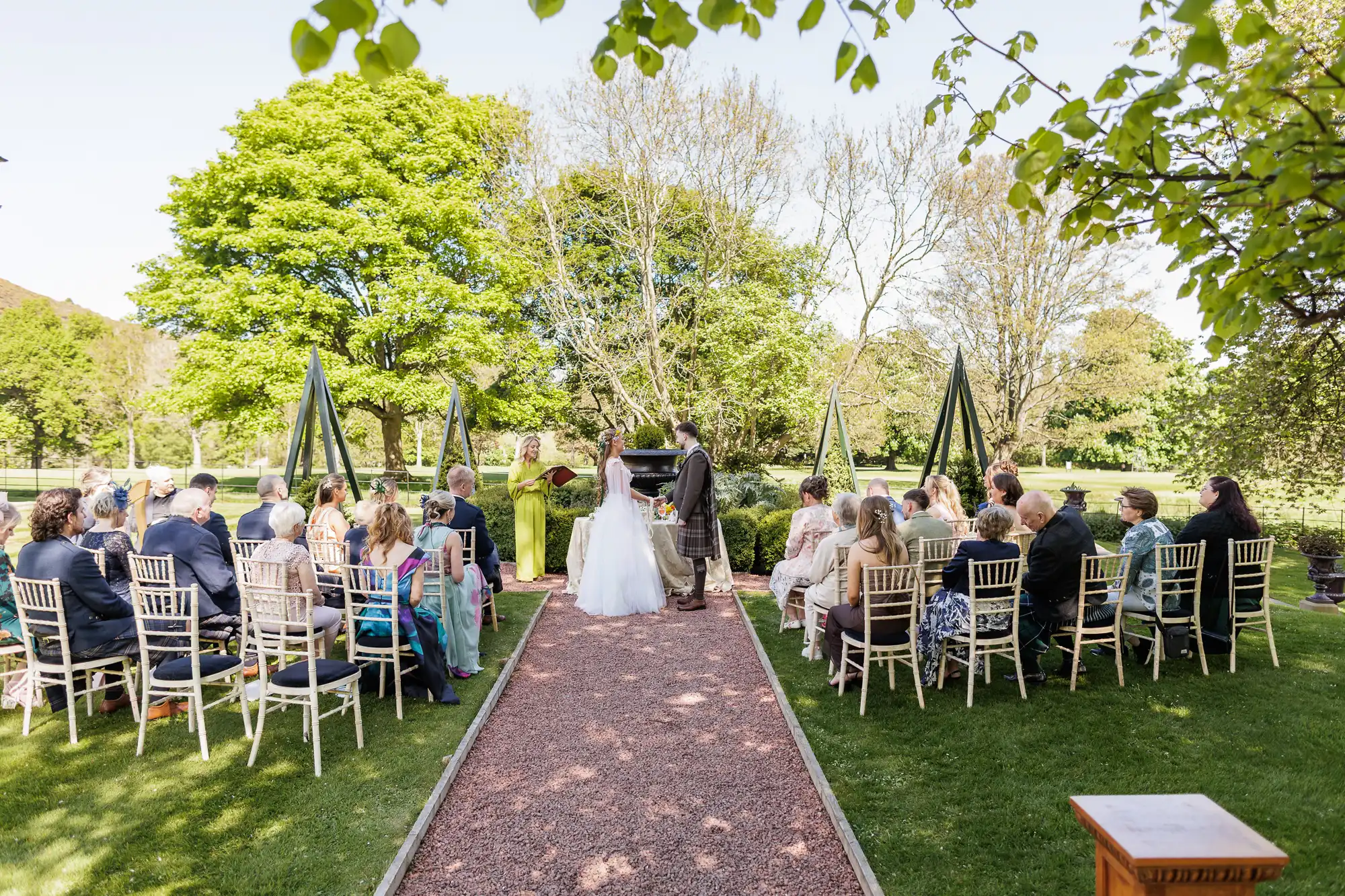 An outdoor wedding ceremony shows a bride and groom standing before an officiant, surrounded by guests seated in rows of white chairs on either side of a path, with green trees and grass under clear, sunny skies.