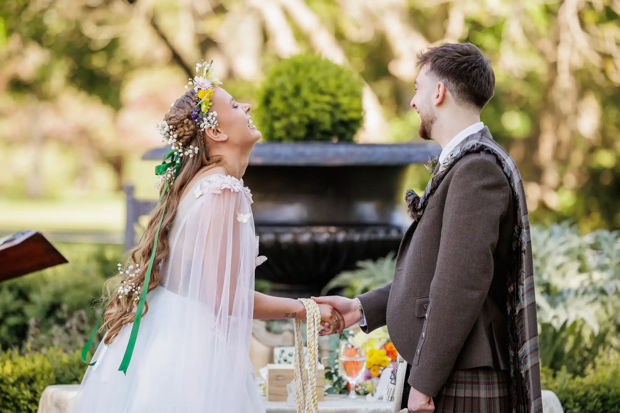 A bride in a white dress and floral crown laughs joyfully while holding hands with a groom in a brown suit and kilt. They stand outdoors in front of a black fountain, surrounded by greenery, with a decorated table nearby.