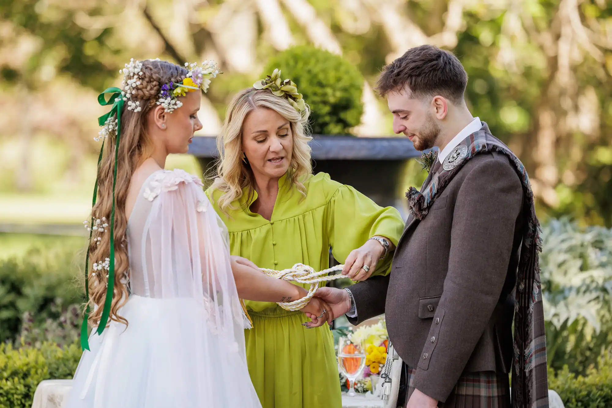 A woman in a green dress ties a cord around the joined hands of a bride and groom during an outdoor handfasting ceremony. The bride wears a white dress and floral crown; the groom wears a suit with a tartan sash. Lush greenery and sunlight create a serene backdrop.