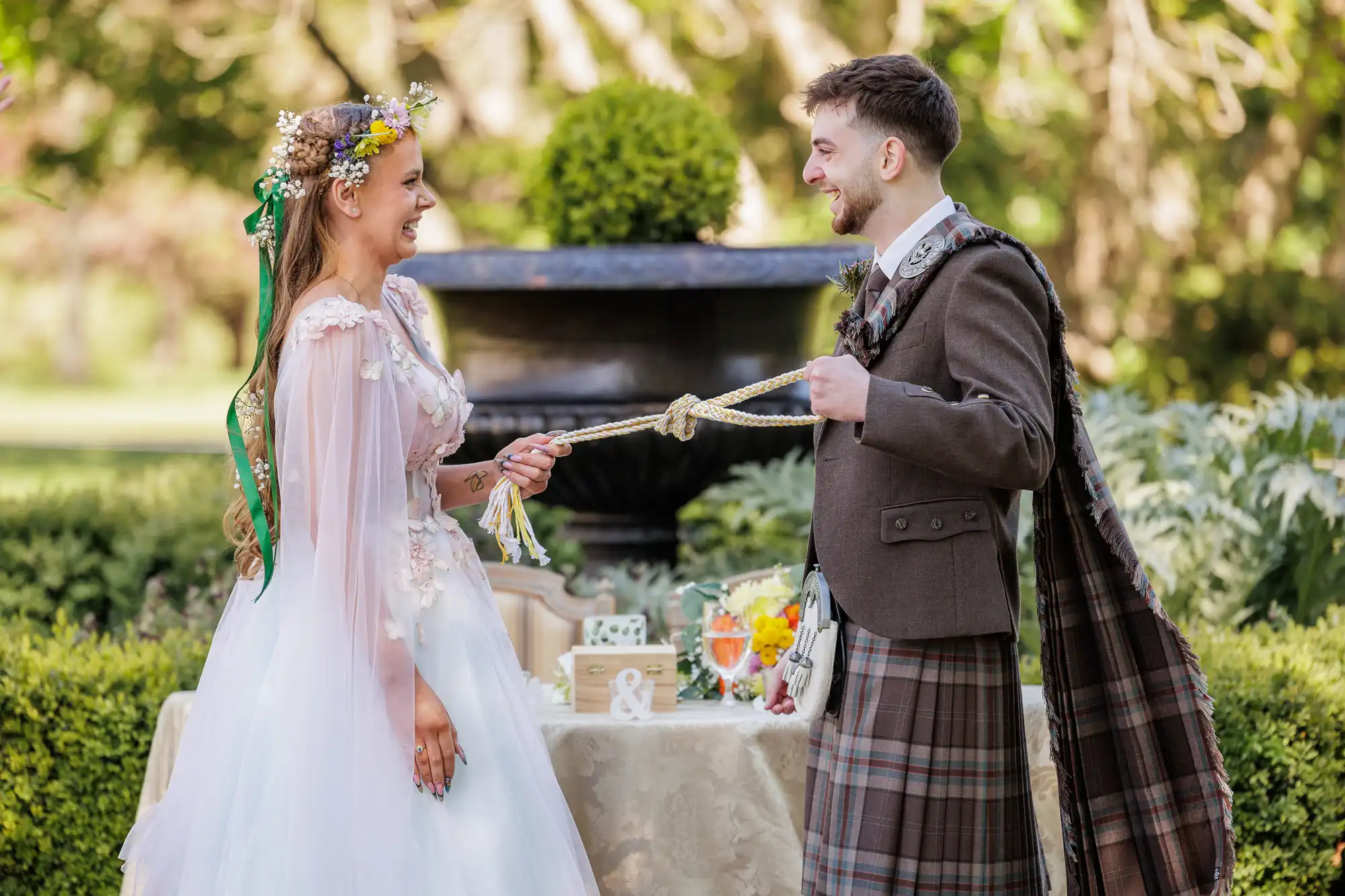 A smiling bride in a white dress with a floral crown and a groom in a kilt perform a handfasting ceremony outdoors, tying their hands with a cord. A table with flowers, drinks, and wedding decor sits behind them in a green garden setting.