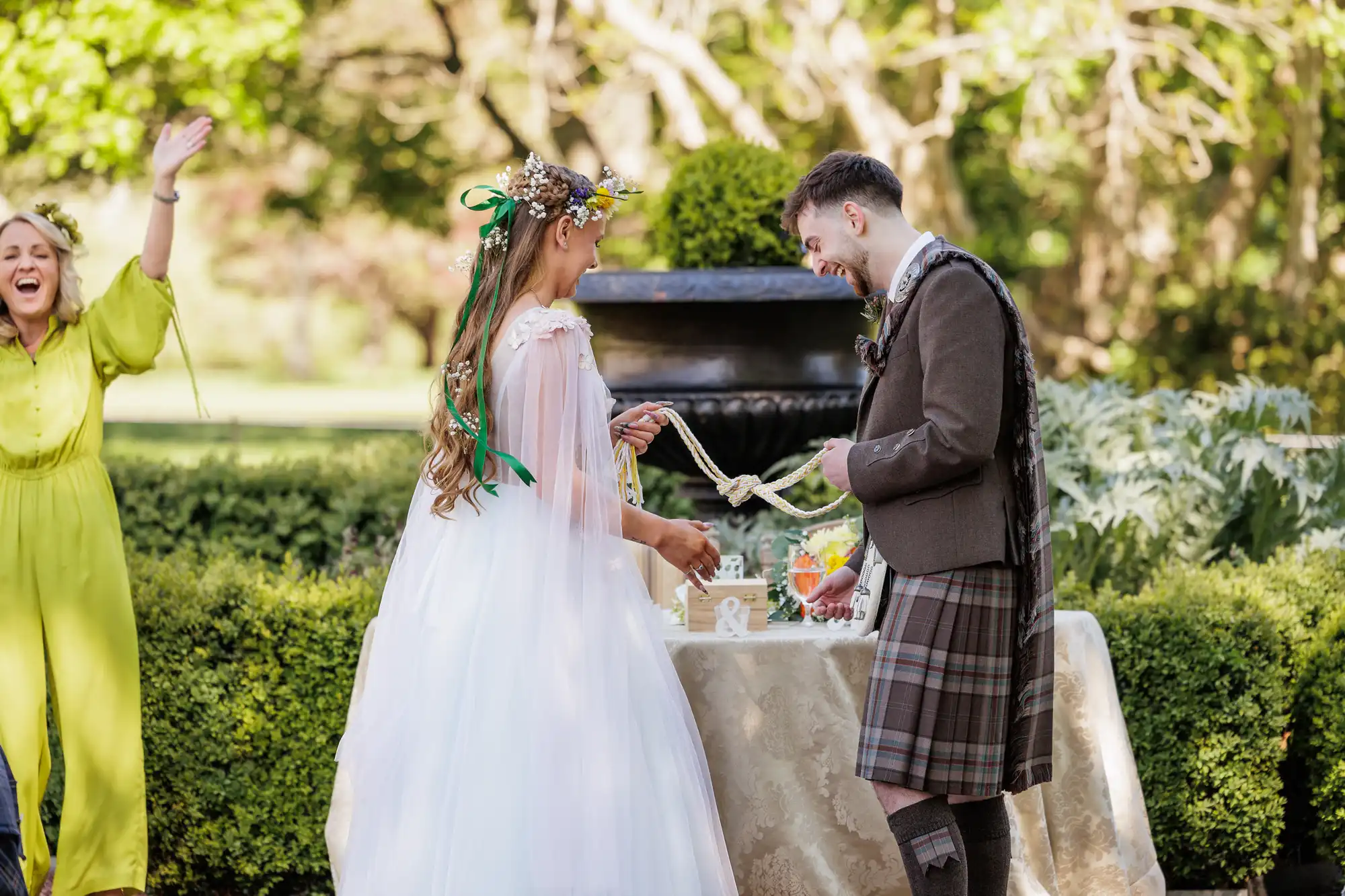 A bride in a white gown and a groom in a kilt smile as they perform a handfasting ritual outdoors. A joyful woman in a yellow dress stands nearby with arms raised. The scene is bright, with greenery and sunlight in the background.