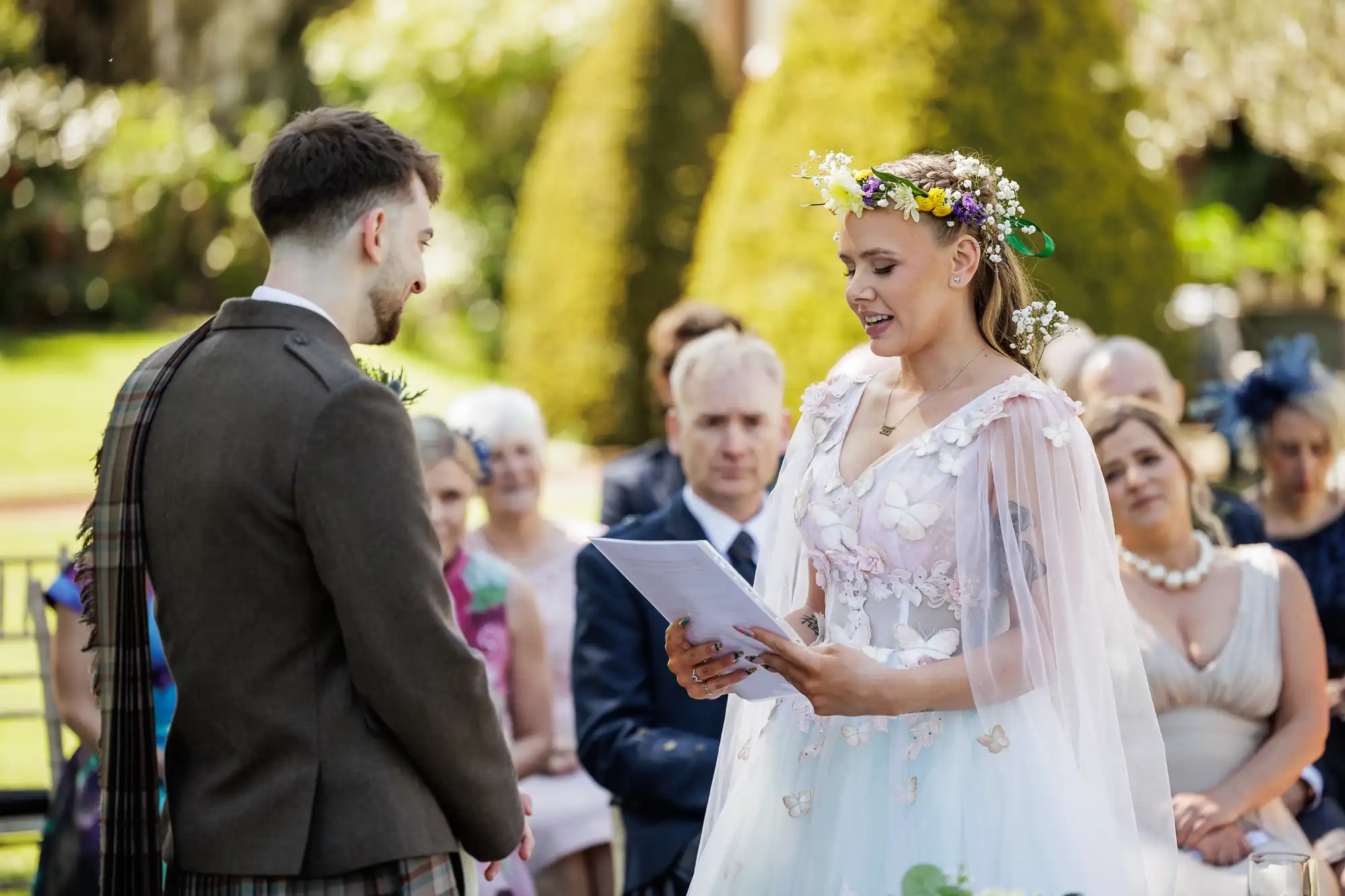 A bride wearing a floral crown and white dress reads from papers to the groom at an outdoor wedding ceremony. Guests in colourful attire sit behind them, attentively watching. Trees and greenery form the background, suggesting a spring or summer setting.