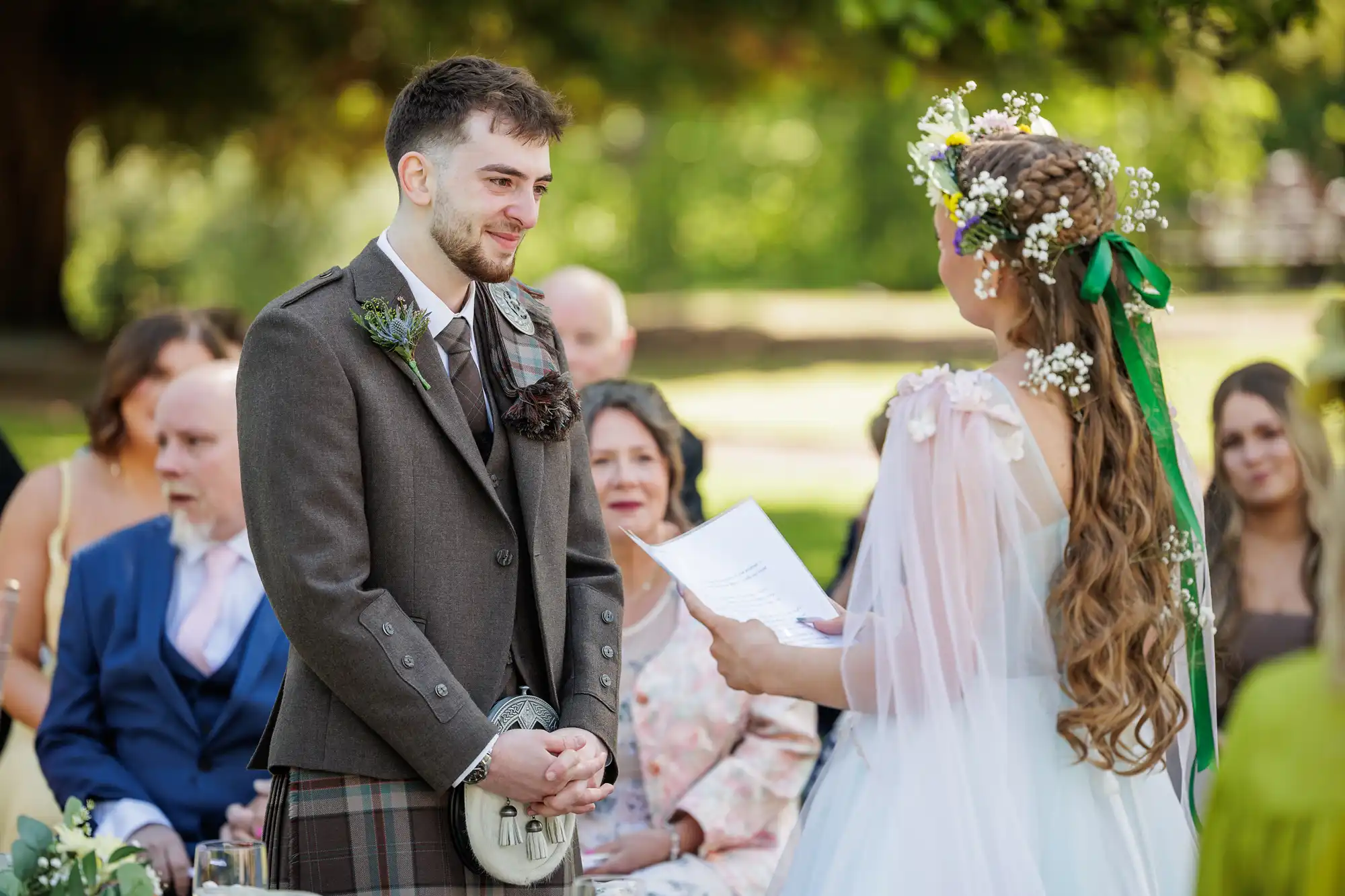 A bride in a floral crown and white dress reads vows to a smiling groom in a brown kilt suit at an outdoor wedding ceremony. Guests sit and watch in the background under green trees, bathed in sunlight.