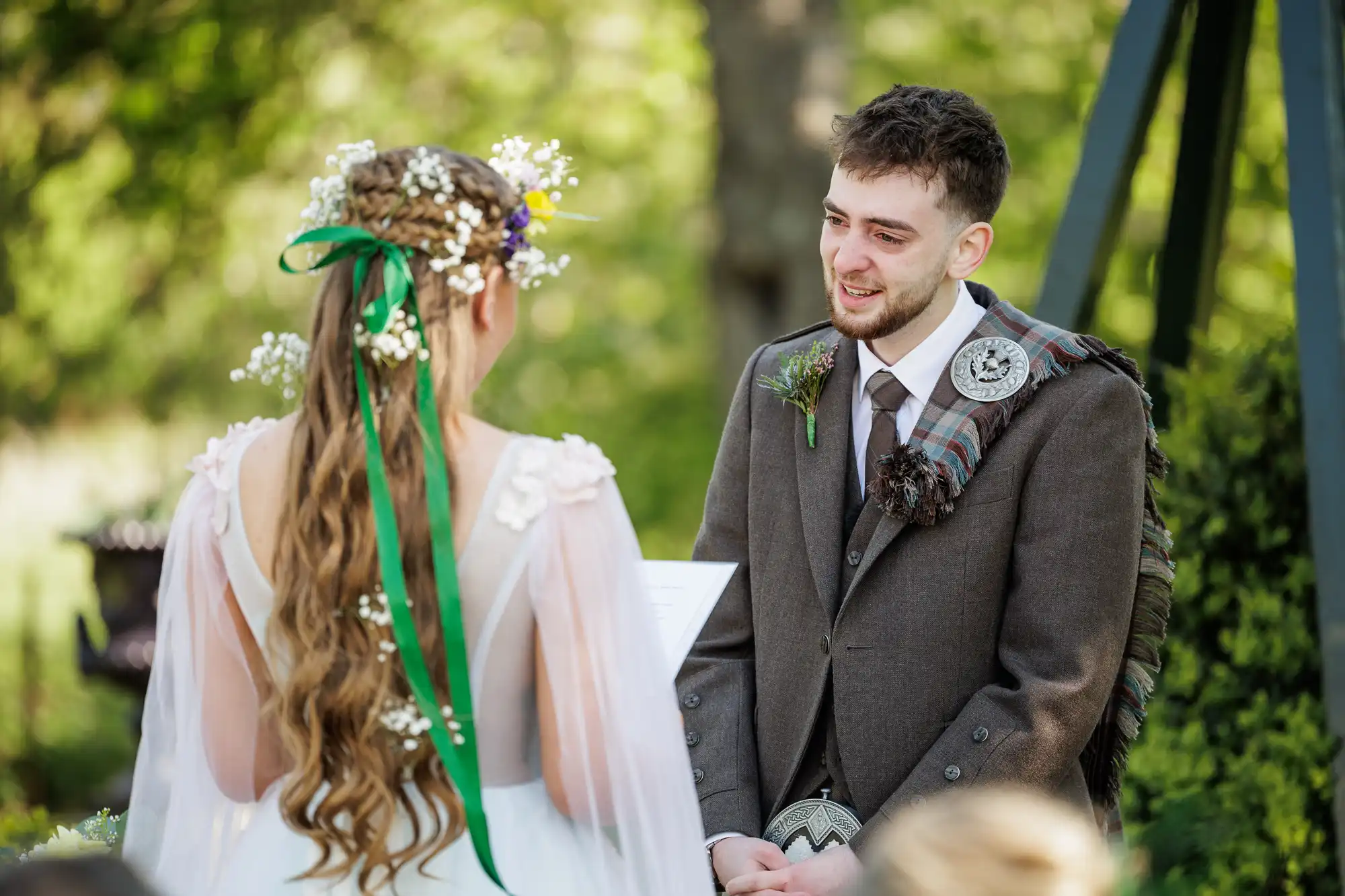 A groom in a brown suit with a tartan sash and buttonhole smiles at a bride in a white dress with floral details and long wavy hair adorned with flowers and green ribbons. They are outdoors, surrounded by greenery, during their wedding ceremony.