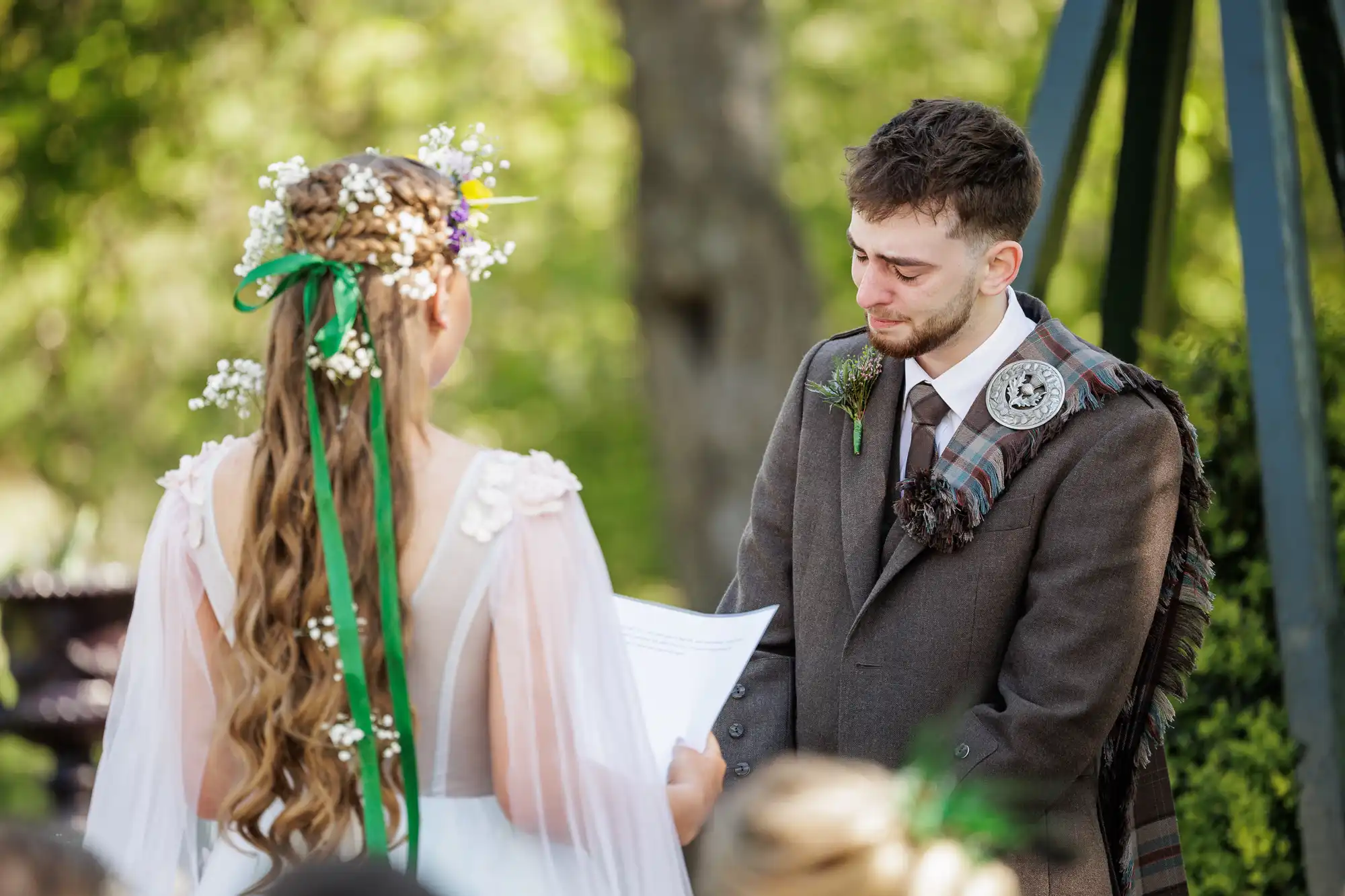 A bride with long, wavy hair and flowers reads vows outdoors to a groom in a brown suit and tartan sash. The groom looks emotional and teary-eyed. Sunlight filters through green trees in the background.
