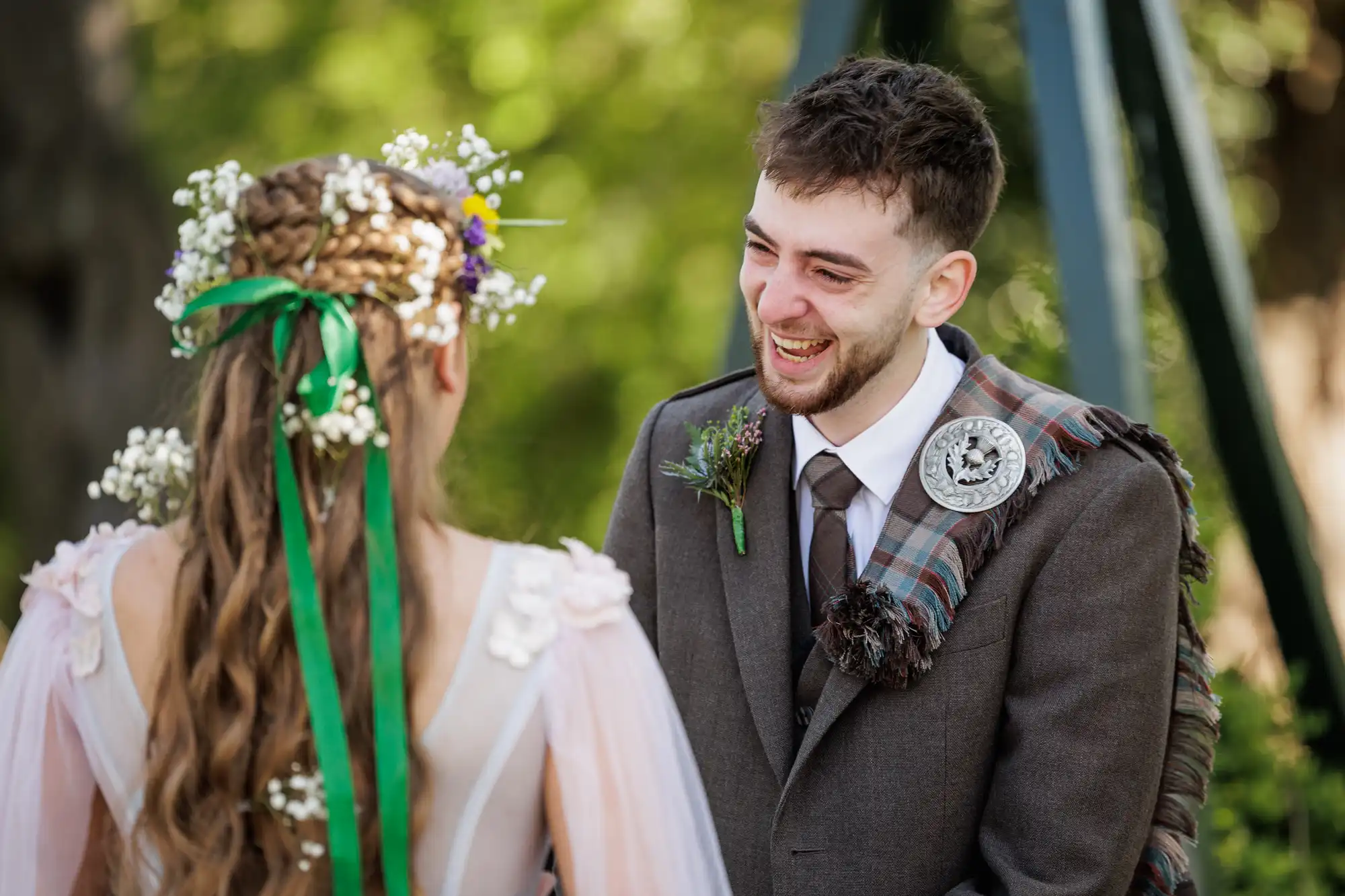 A smiling man in a suit with a tartan sash and brooch faces a woman with long plaited hair, adorned with flowers and ribbons, wearing a dress with sheer sleeves. They are outdoors with green foliage blurred in the background, sharing a joyful moment.