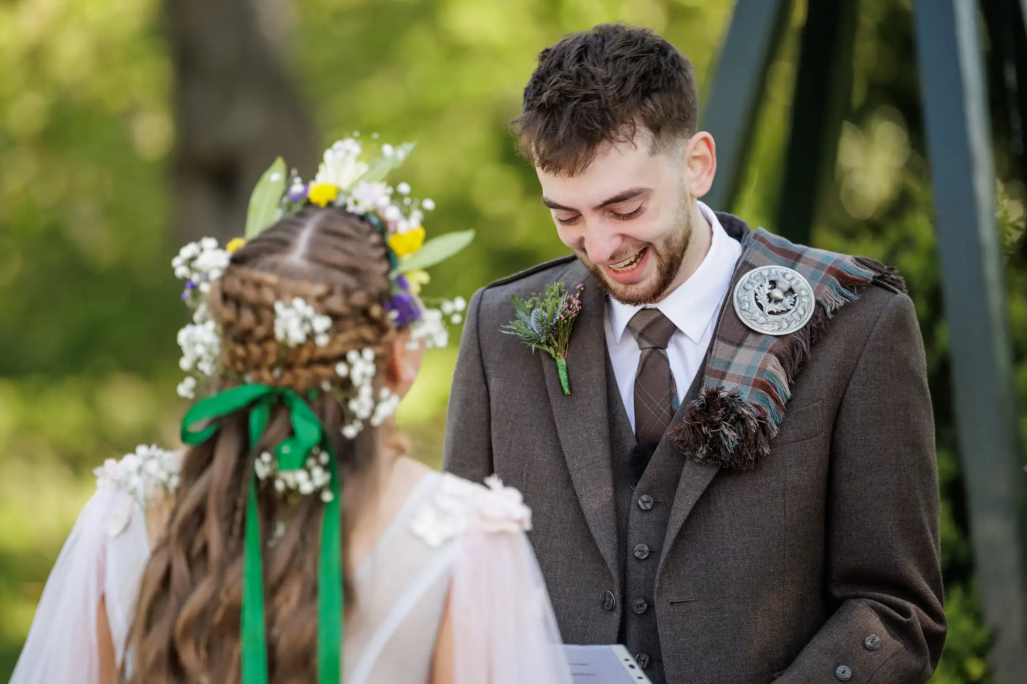 A groom in a brown suit with a tartan sash and buttonhole smiles warmly at a bride with plaited hair decorated with flowers and green ribbons; they stand outdoors amid greenery, exchanging vows or speaking during a wedding ceremony.