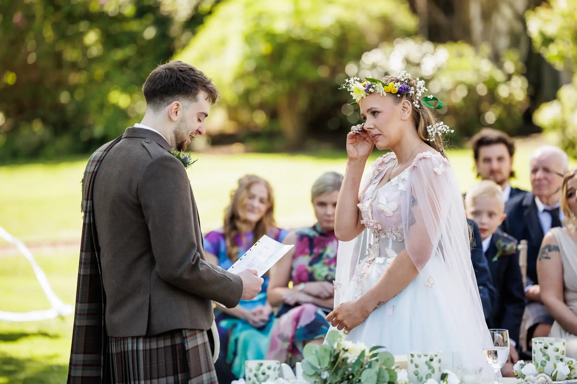 A groom in a brown kilt reads vows to a bride in a white dress and floral crown, who wipes a tear from her eye. Guests sit behind them outdoors, watching the emotional moment amid greenery and sunlight. Flowers and tea sets decorate the table in the foreground.