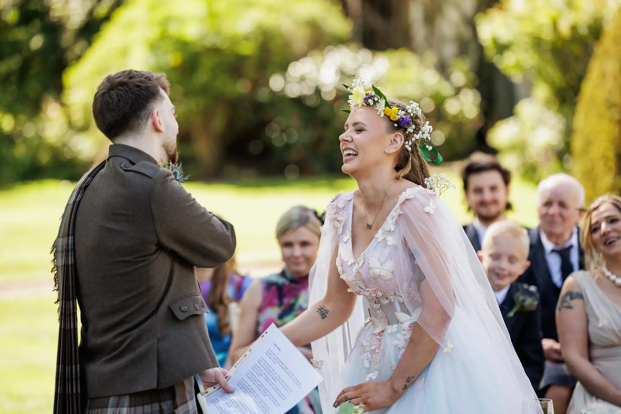 A bride in a floral crown and white dress laughs joyfully, holding papers, during an outdoor wedding ceremony. The groom, wearing a brown suit, faces her. Guests, seated in colourful attire, watch and smile in a lush garden setting.