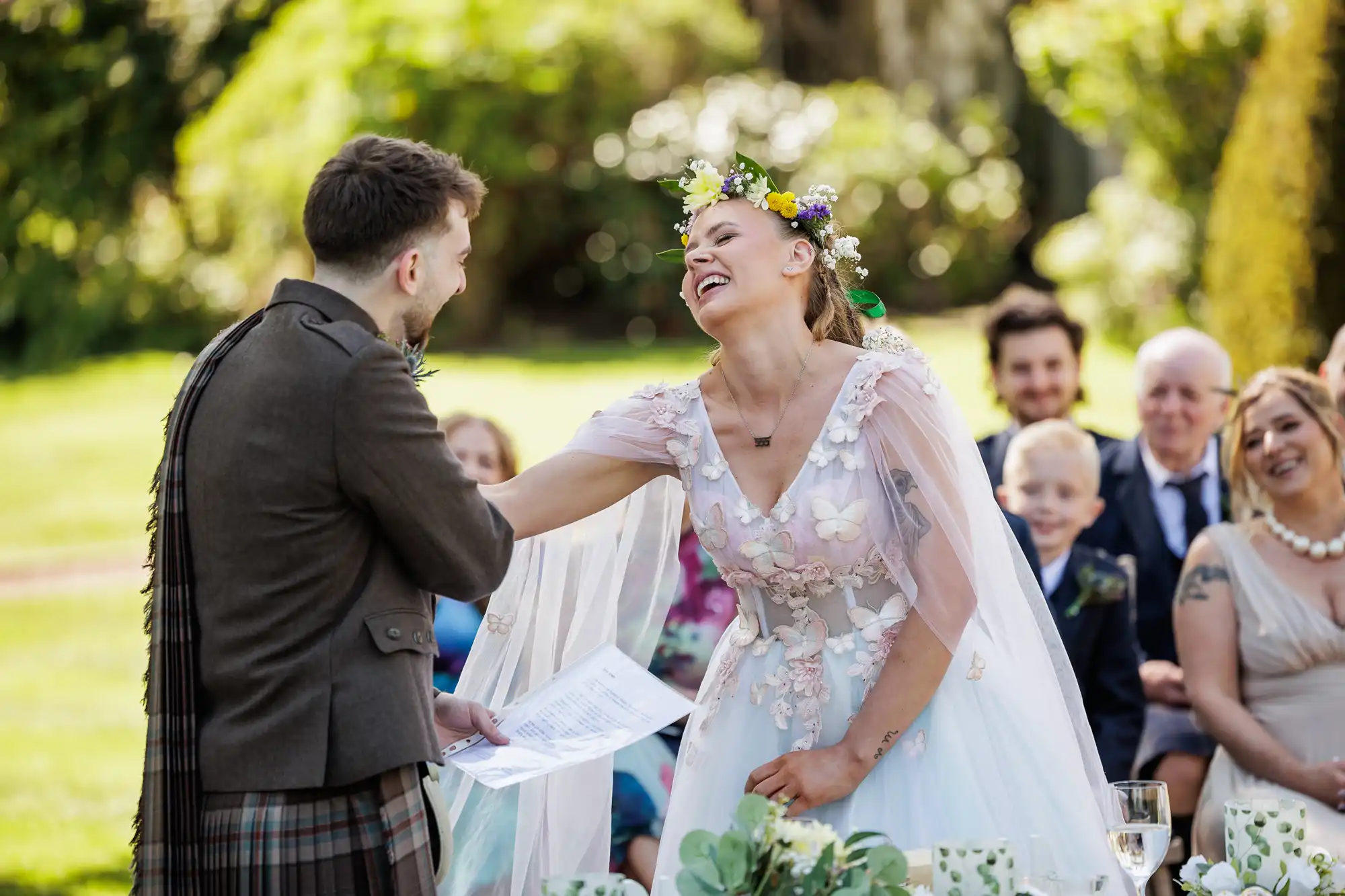 A bride in a floral crown and white gown laughs joyfully whilst shaking hands with the groom, who wears a brown jacket and kilt. Guests smile in the background at the outdoor wedding, surrounded by greenery and sunlight. The bride holds papers, possibly vows.