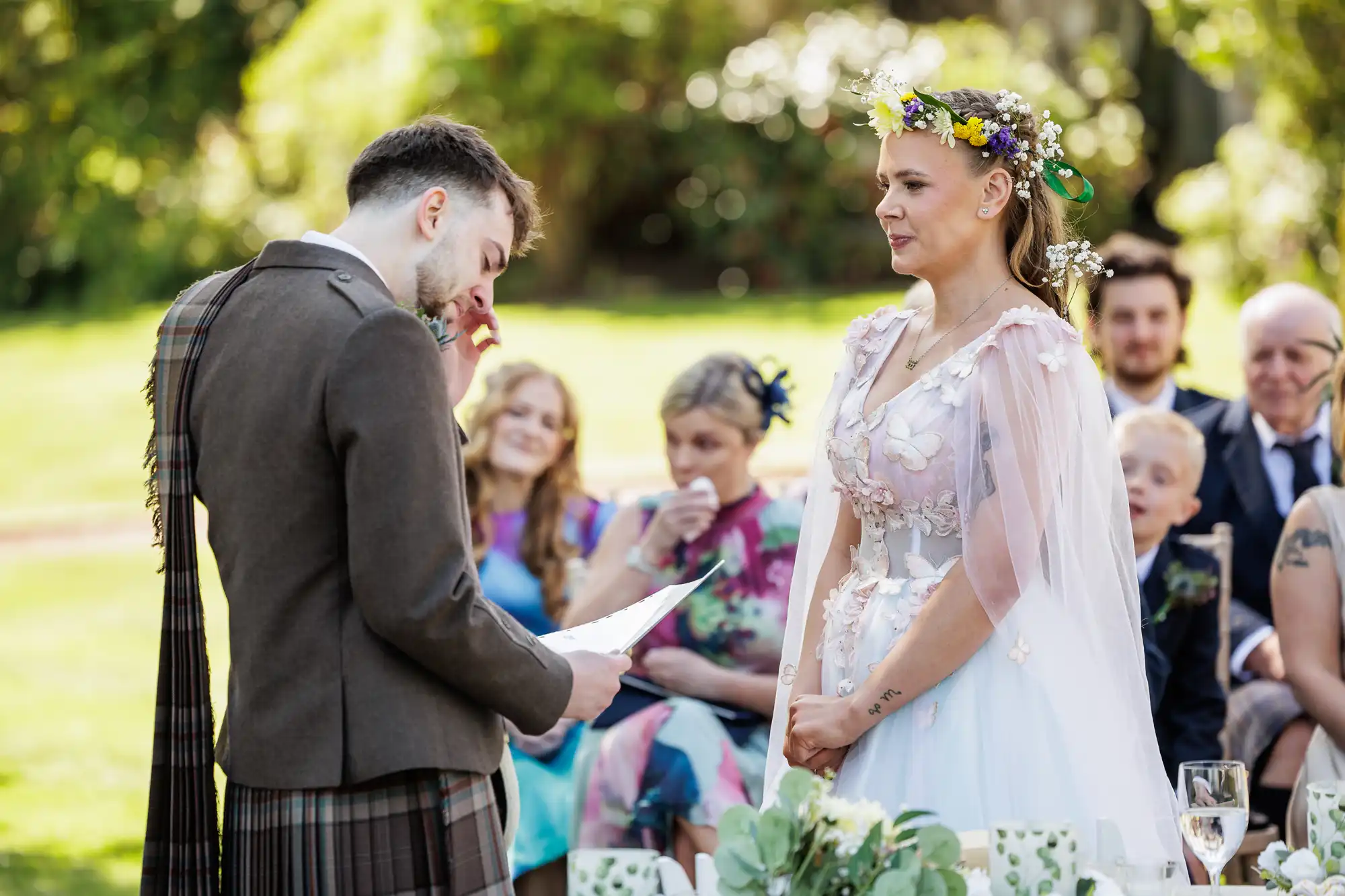 A groom in a brown kilt reads vows while wiping his eye; a bride in a floral crown and white dress listens, hands clasped. Guests, some emotional, watch in the background at this outdoor wedding ceremony on a sunny day.