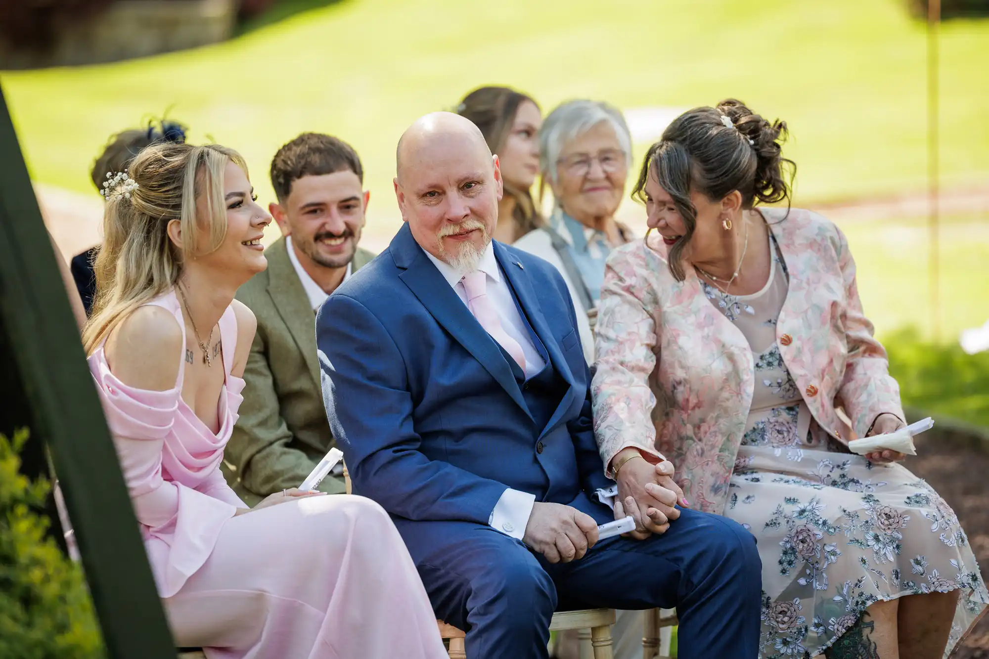 A group of people sit outdoors at a formal event. A man in a blue suit and pink tie sits in the centre, holding hands and smiling with a woman in a floral dress. A young woman in a light pink dress and others in formal attire sit nearby, all smiling and laughing.