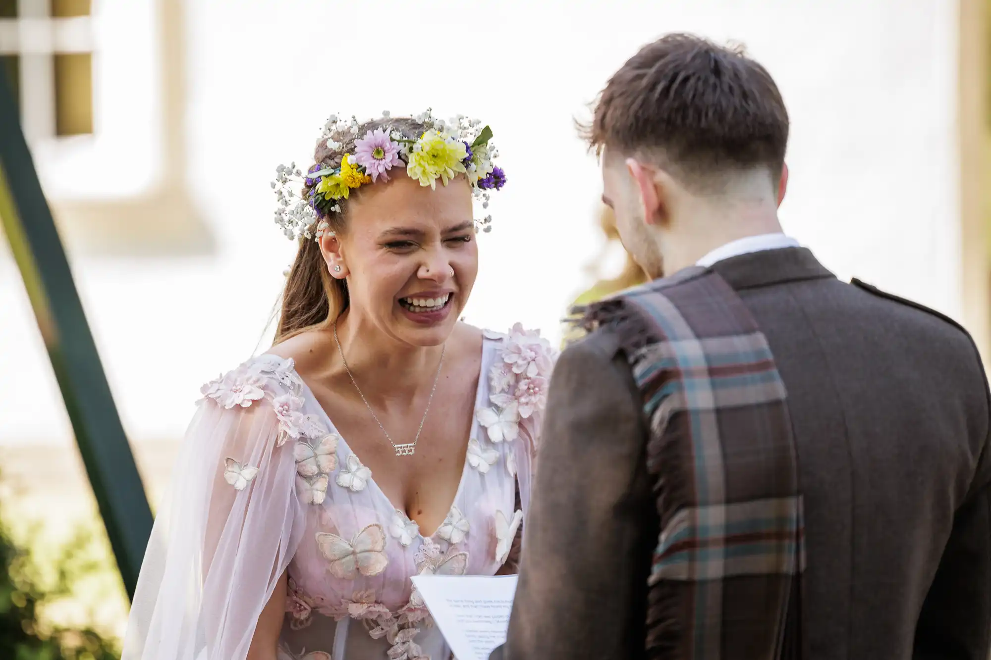 A smiling bride wearing a flower crown and a white dress with floral and butterfly details faces a groom in a brown suit with a tartan sash. The bride appears joyful during an outdoor ceremony, holding her vows while sunlight highlights the scene.