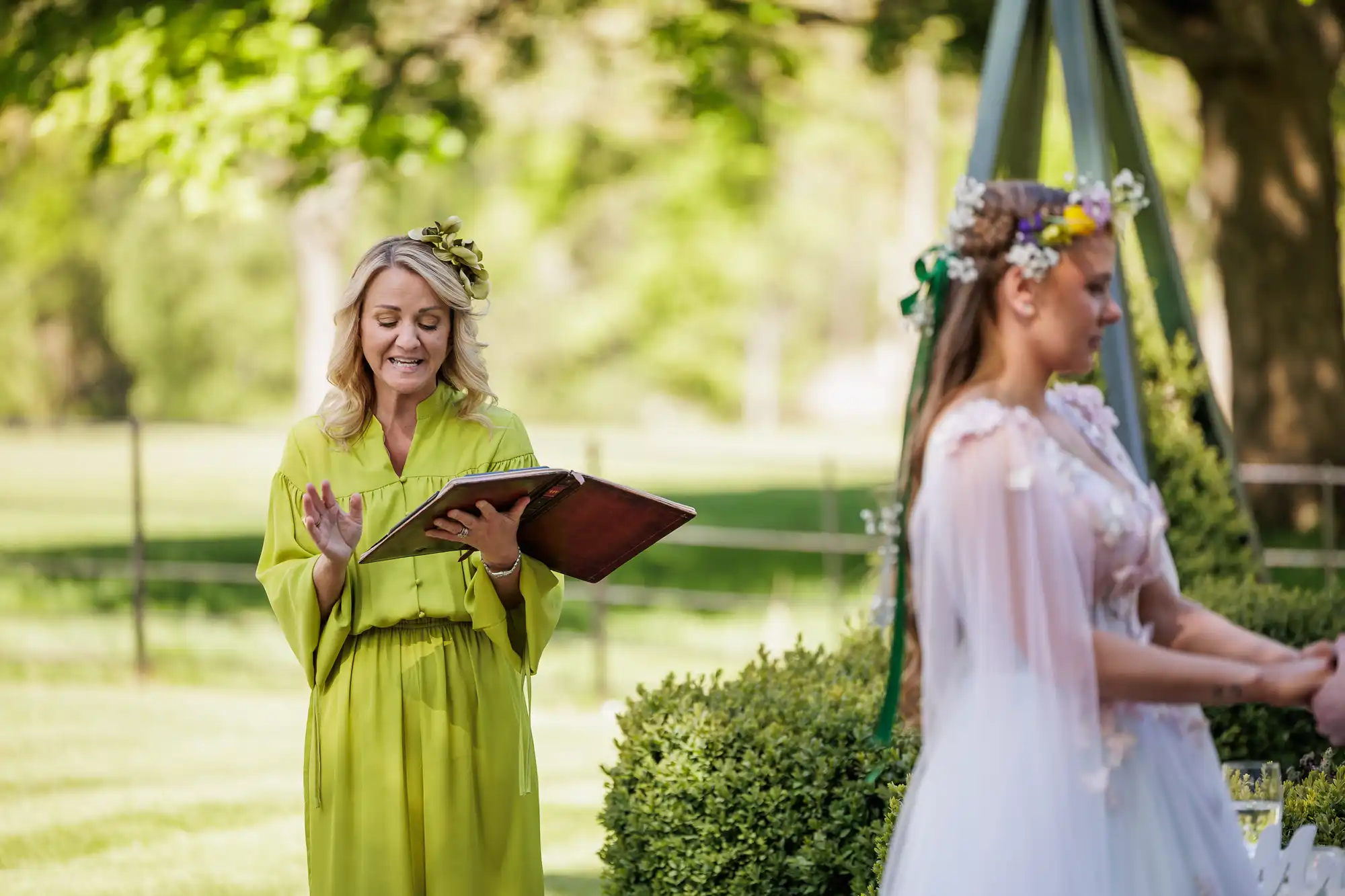 A woman in a green dress and matching hat joyfully reads from an open book outdoors. Next to her, a bride in a white dress with a flower crown stands, hands clasped, during a garden ceremony with greenery and sunlight in the background.
